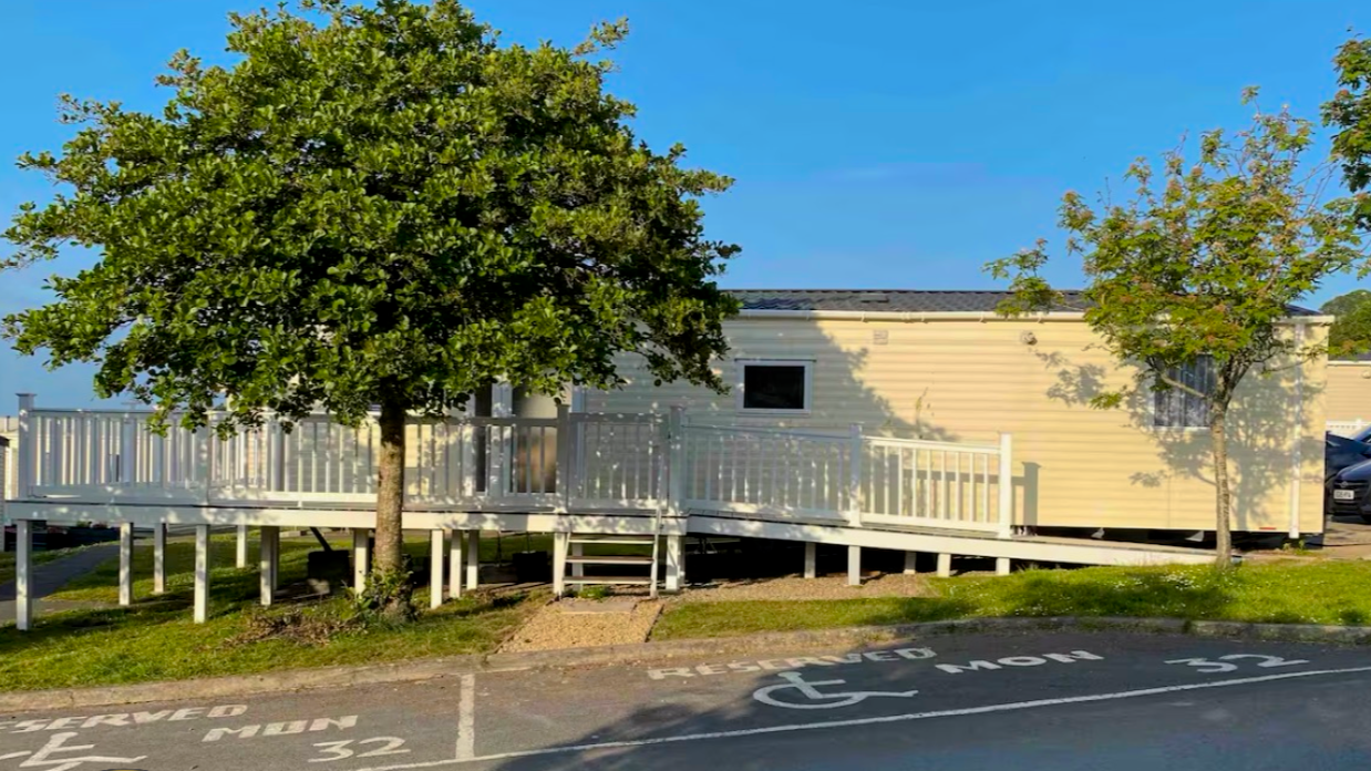 A beige mobile home with a black roof, white railing, and a small set of stairs leading to the entrance, situated behind two trees on a grassy area next to a parking space.