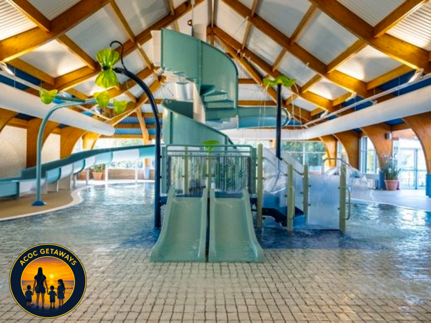 Indoor water park with a large enclosed pool, water slides, and a high wooden ceiling with exposed beams. The pool has a textured tile floor, and the water slides are in pastel colors. There are potted plants and large windows allowing natural light.