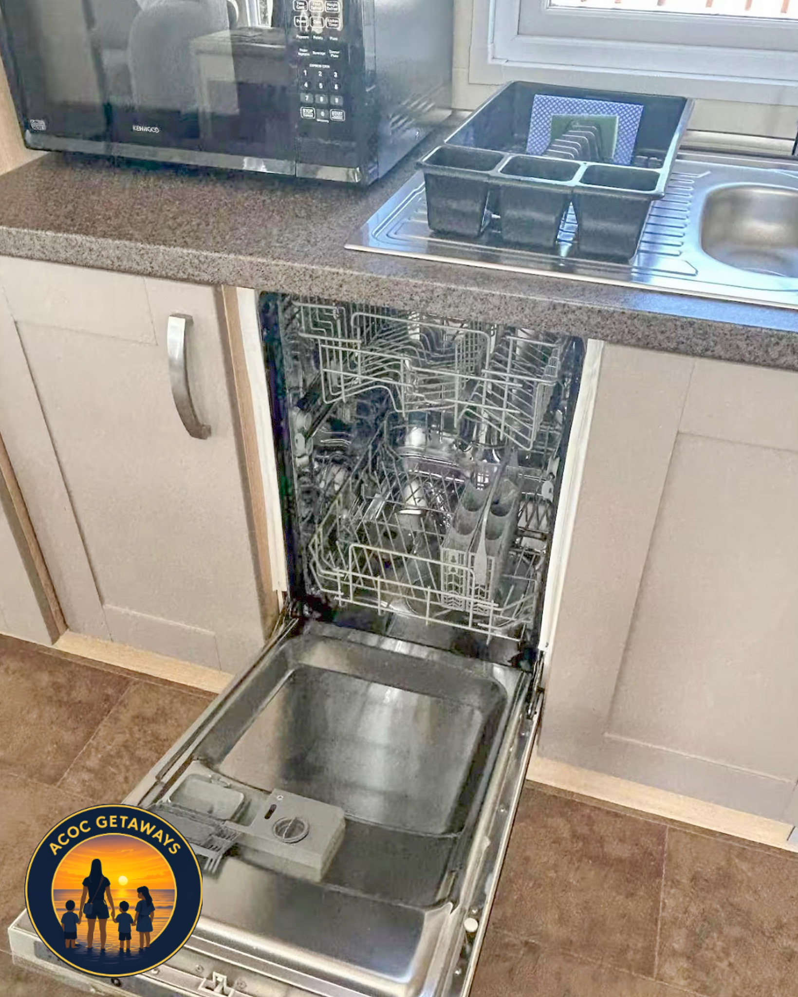 The image shows an empty dishwasher with the door open, placed beneath a kitchen countertop. There are compartments for utensils and a tray inside, and it is within a kitchen area with a tiled floor.