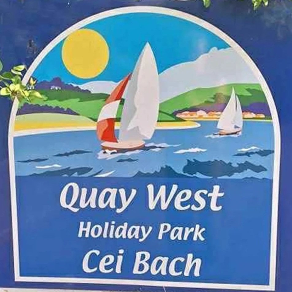 Sign for Quay West Holiday Park, featuring a beach scene with sailboats, green hills, a bright yellow sun, and blue water.