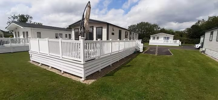 View of a white wooden deck with a railing attached to a small house, surrounded by a grassy yard and other similar houses nearby.