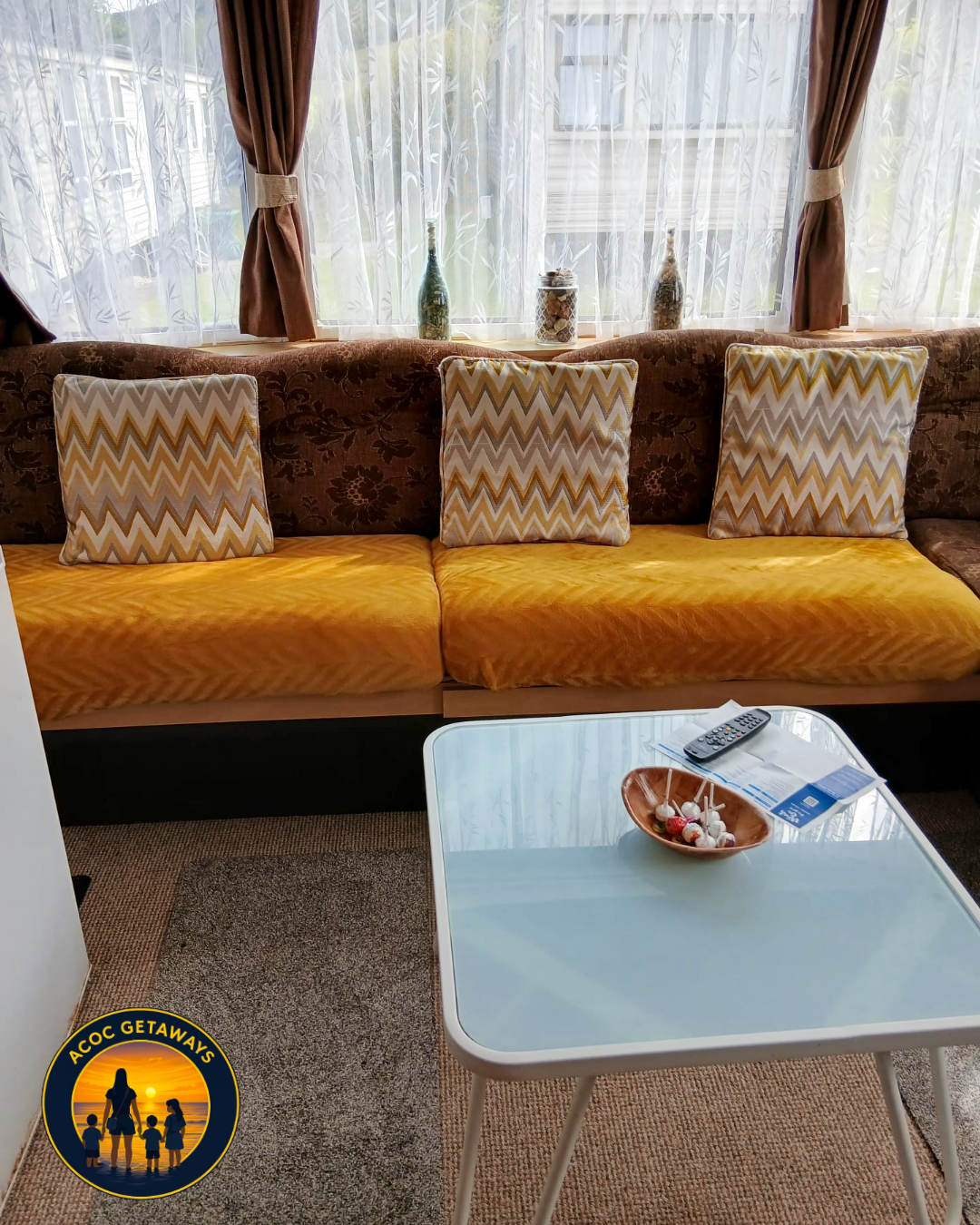 Living room with a brown floral couch with yellow cushions and three decorative pillows, a window with curtains, a white coffee table with a bowl of lollipops, a remote control, and some papers. The room has beige carpet flooring.