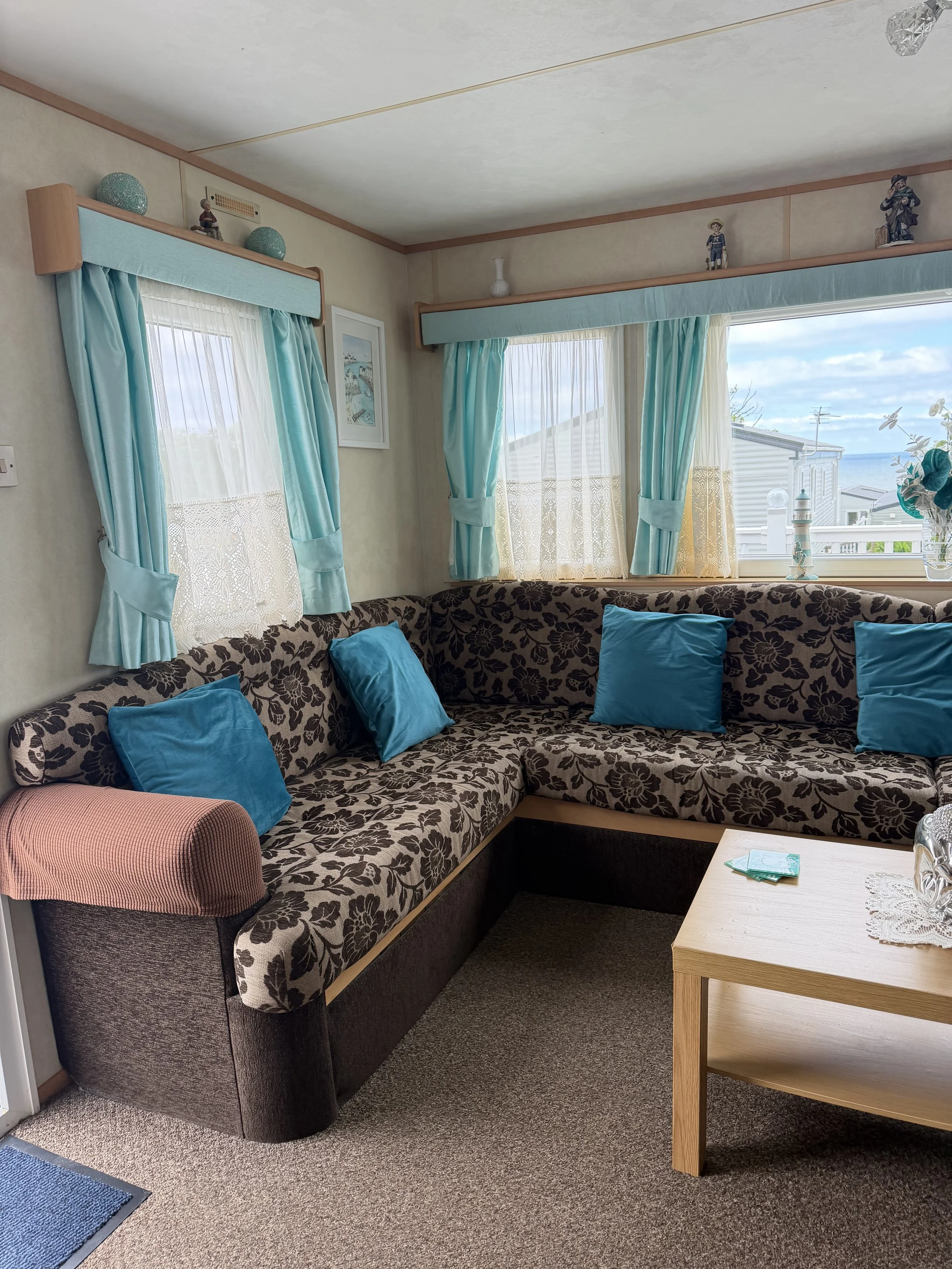 Living room with patterned sofa, blue pillows, wooden side table, windows with light blue curtains, decorative figurines on window ledge, and ocean view outside.