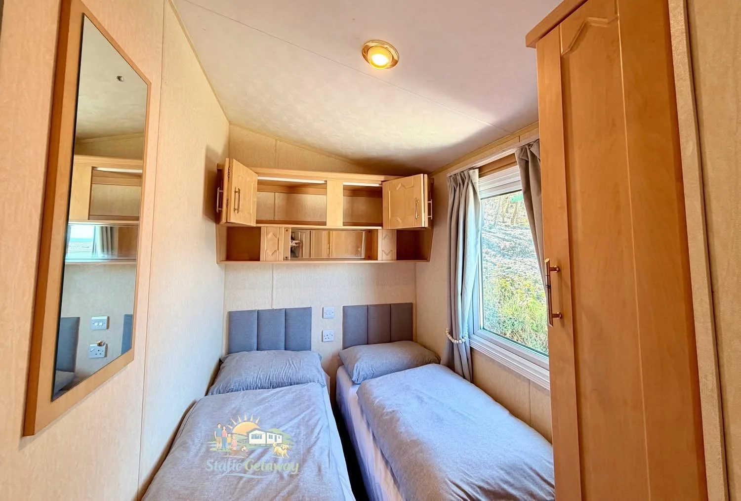 Interior of a small bedroom with two single beds with gray headboards, dressed in beige bedding, next to a large window with beige curtains. There are wooden cabinets above the beds, a wooden wardrobe on the right, and a mirror on the left wall.