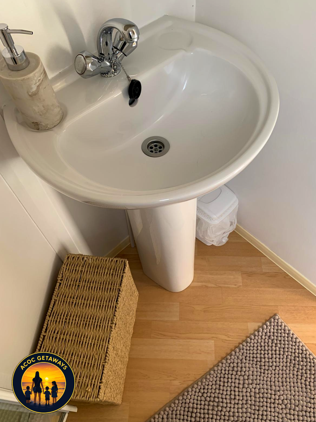 Bathroom sink with soap dispenser, small trash bin, woven storage box, and textured gray rug on wooden floor.