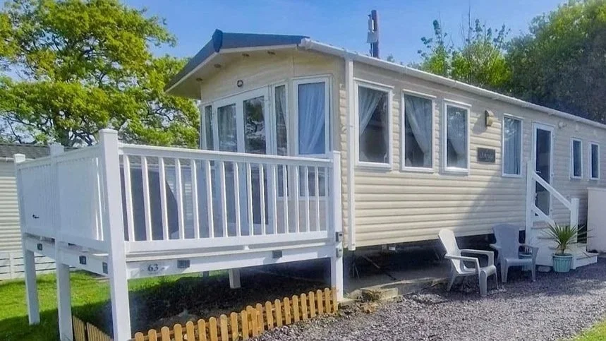 A beige mobile home with multiple windows, a small front porch with white railing, and outdoor chairs. There are trees and a clear blue sky in the background.