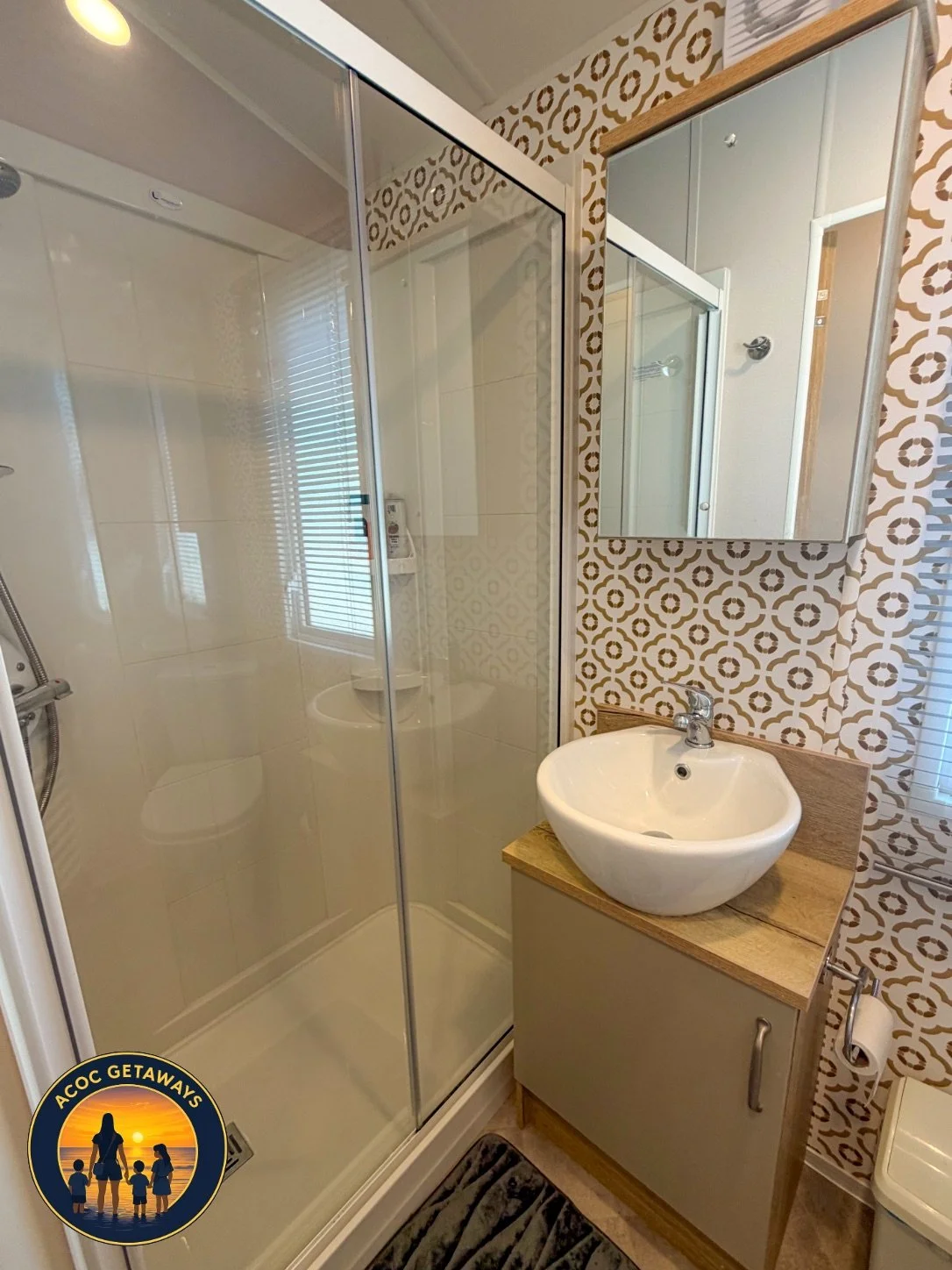Small bathroom with a glass shower enclosure, a white round vessel sink on a wooden vanity, a wall-mounted mirrored medicine cabinet, patterned wallpaper, and a window with blinds.