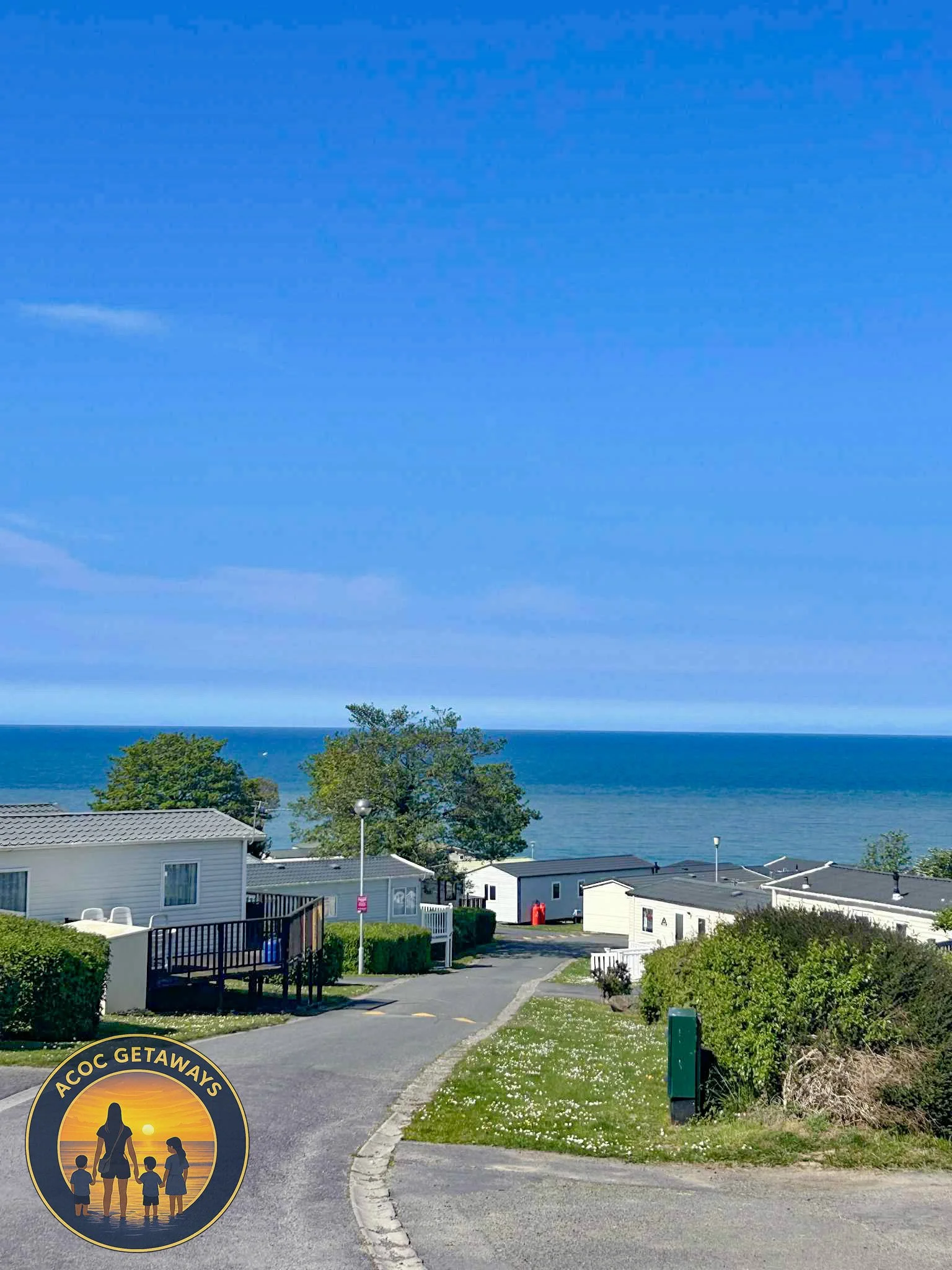 View of a coastal holiday park with white caravans, a paved road, green bushes, and trees, overlooking the ocean under a clear blue sky.