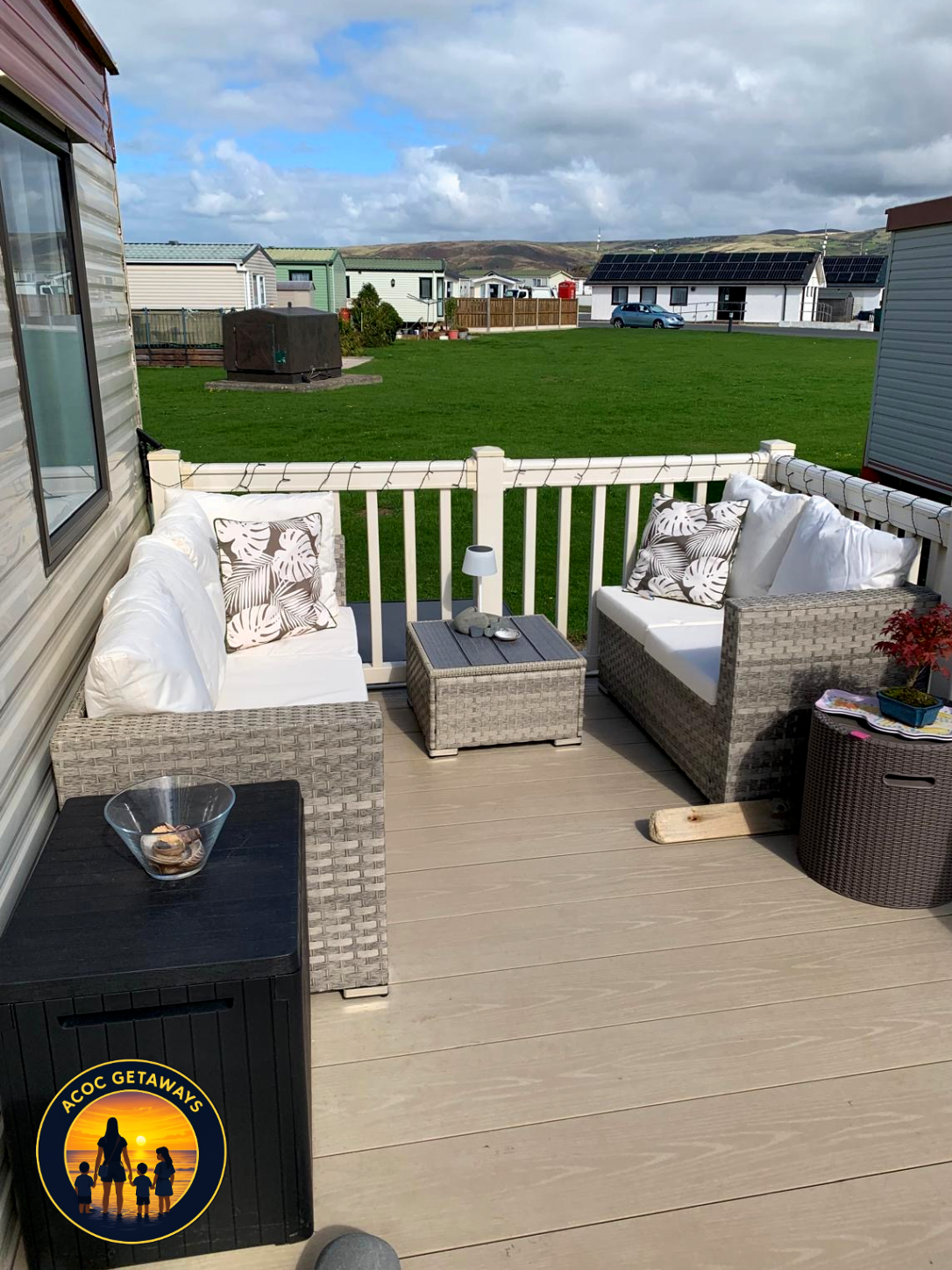 Cozy outdoor patio with two wicker sofas with white cushions and patterned throw pillows, a small side table with a lamp and stones, on a wooden deck overlooking a grassy backyard and neighboring houses with solar panels.