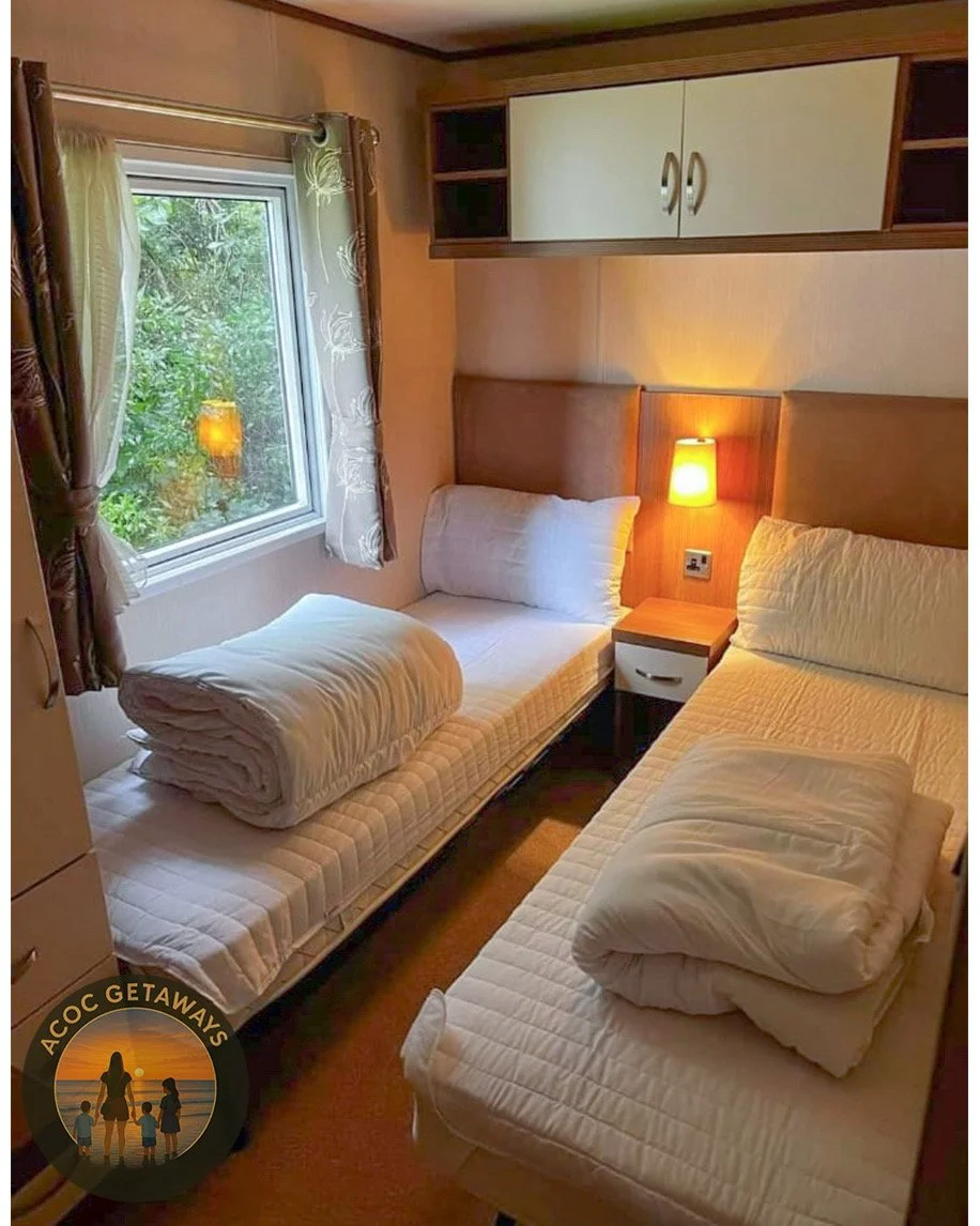 A cozy bedroom with two single beds, each with rolled-up white bedding on top. A small nightstand with a lamp is positioned between the beds, and a window with leafy greenery outside is on the left, framed by patterned curtains. There is a wooden cabinet with storage compartments above the beds.