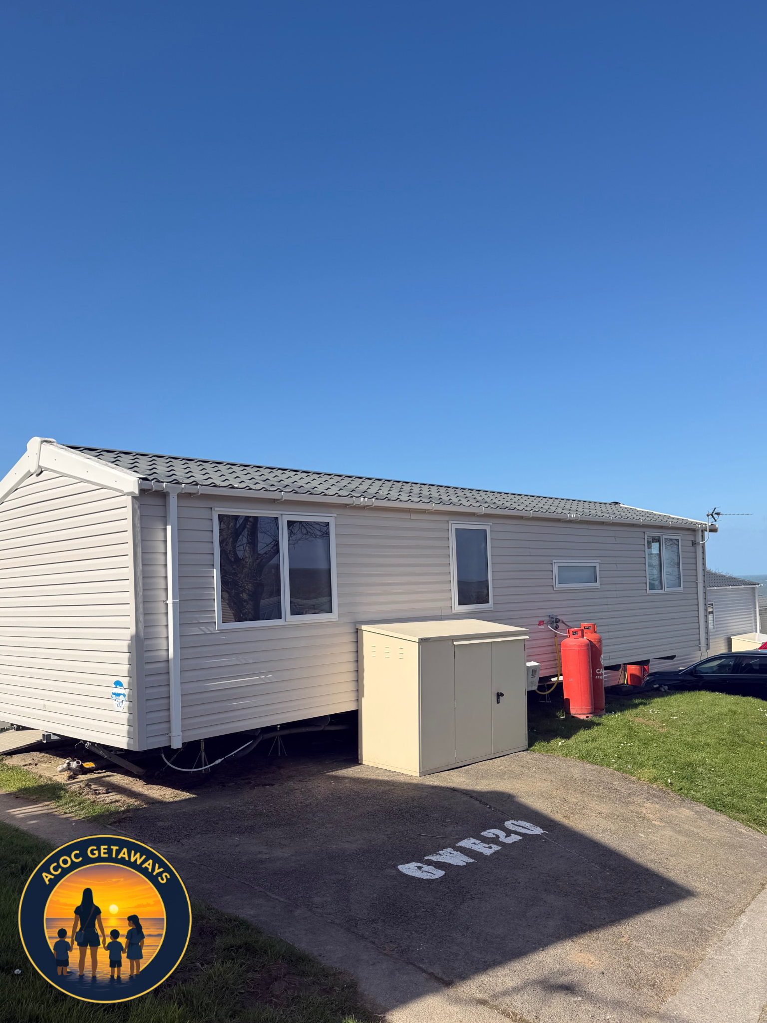 A beige mobile home with three windows, a beige storage shed, and two red propane tanks, situated on a concrete slab with painted 