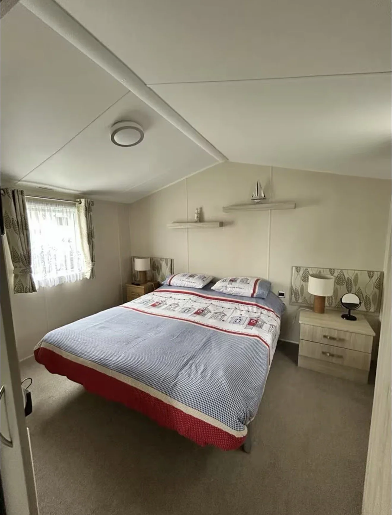 A cozy bedroom with a bed that has a patterned quilt and pillows, two wooden nightstands with lamps, a shelf with decorative items, a window with curtains, and light-colored walls and carpet.