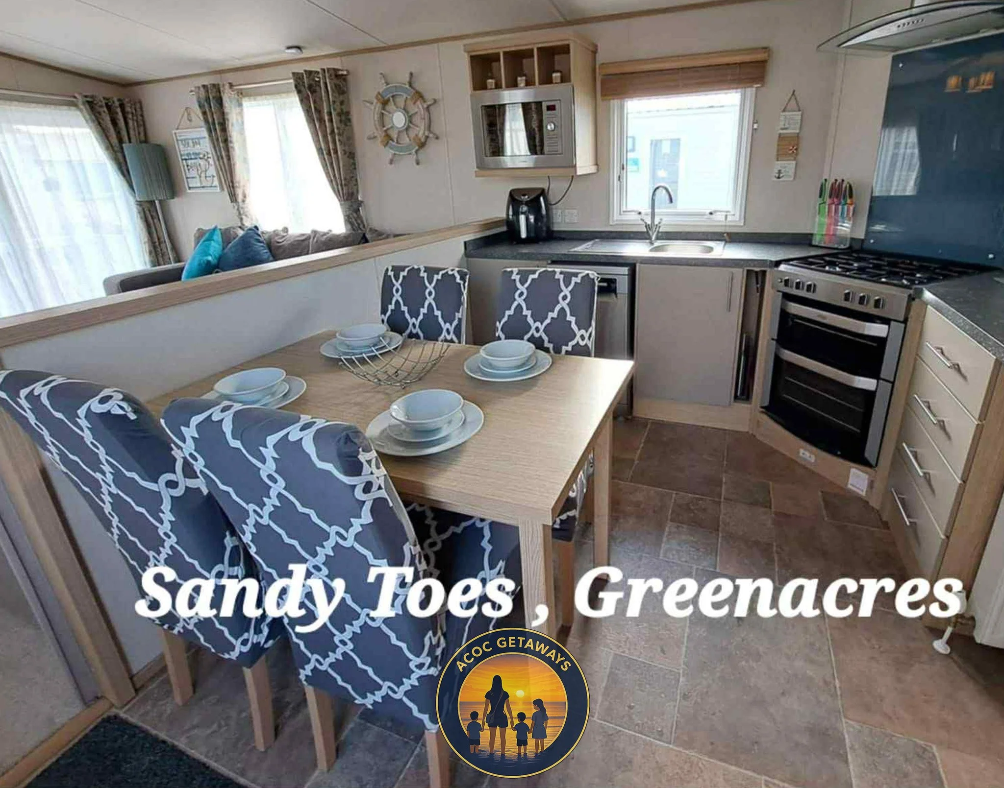 Interior view of a cozy kitchen and dining area with a light wood dining table and four patterned chairs, set with white bowls and plates. The kitchen features a microwave, coffee maker, sink, and stove. Bright windows and nautical decor give a beachside vibe. The text overlay reads 'Sandy Toes, Greenacres' with a logo below showing a family silhouette at sunset with the caption 'ACOC GETAWAYS'.