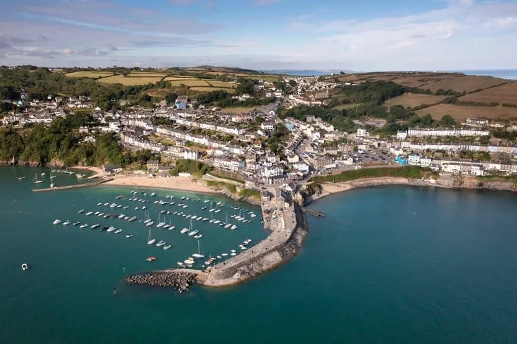 A coastal town with a harbor filled with boats, surrounded by green hills and residential buildings, under a partly cloudy sky.