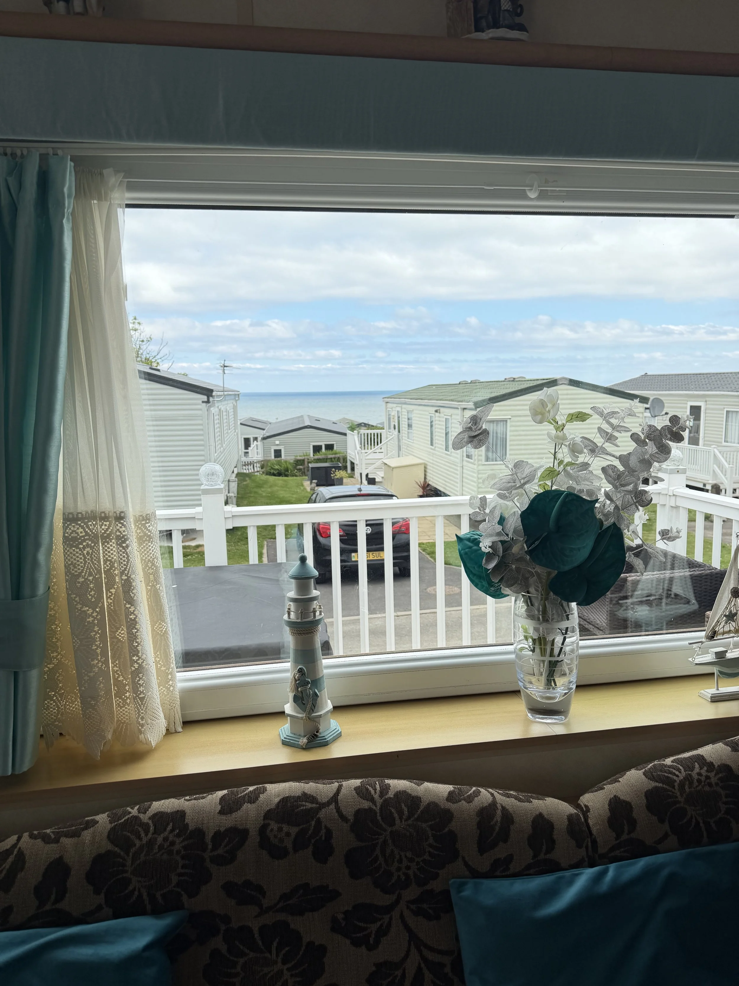 View from a window of a coastal neighborhood with mobile homes, a parked car, a white fence, and a cloudy sky over the ocean.