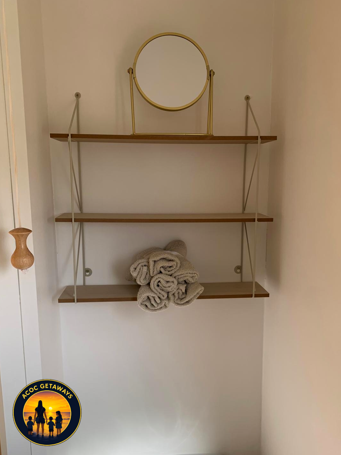 White wall-mounted shelves with a small mirror on top, folded towels on the bottom shelf, and a logo in the bottom left corner reading "ACOC GETAWAYS" with a silhouette of a family by the beach at sunset.