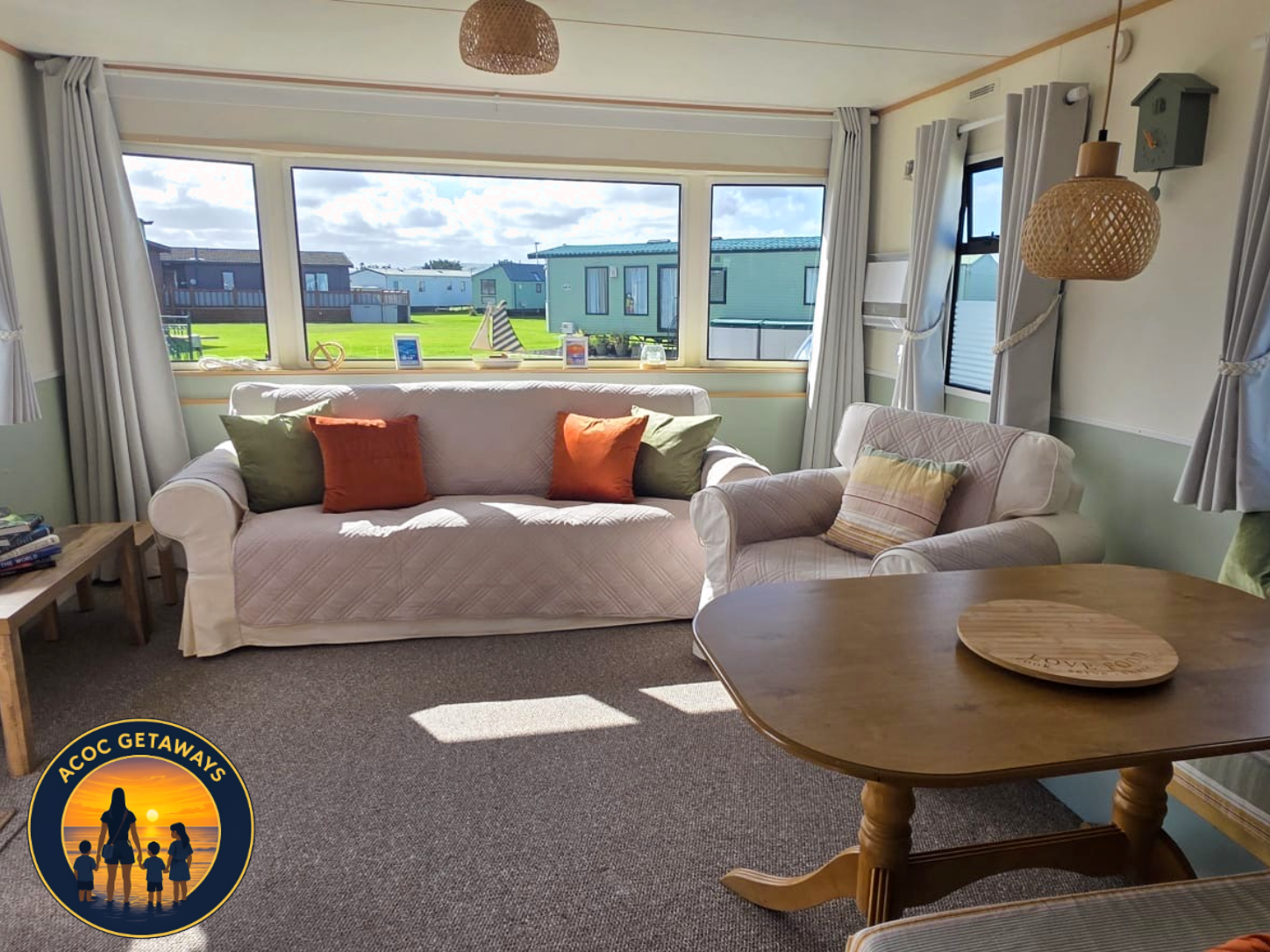Living room with large bay window, white sofas with colorful cushions, wooden side tables, and hanging wicker light fixtures, overlooking a grassy area with mobile homes outside.