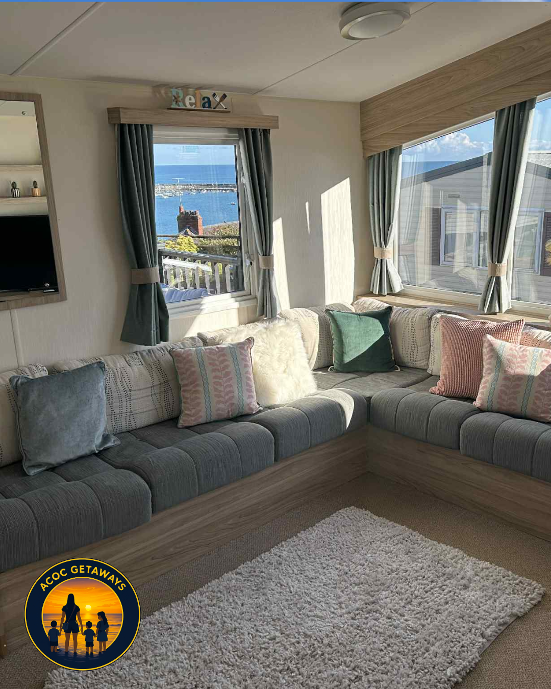 Cozy living room with corner window view of a marina and the ocean, featuring a built-in gray sectional sofa with colorful pillows, curtains, and a white rug.