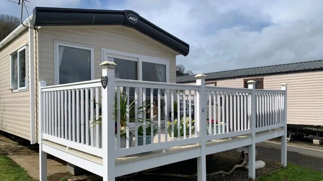 White wooden deck attached to beige mobile home with sliding glass doors and potted plants, surrounded by neighboring mobile homes, under cloudy sky.