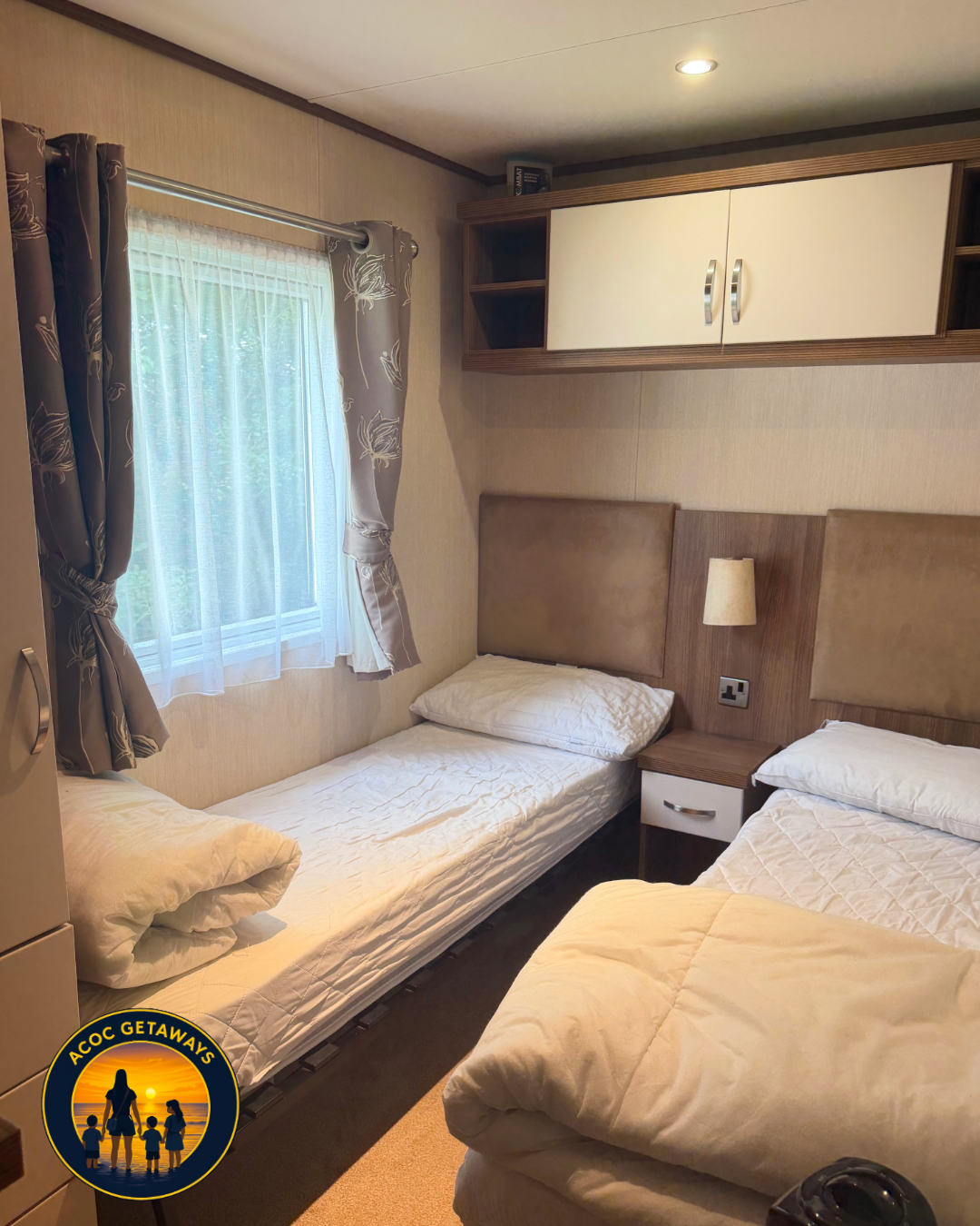 A small bedroom with two single beds, one with a pillow and bedspread, and the other with pillows, a headboard, a small nightstand with a lamp, a window with curtains, and a wall cabinet above.