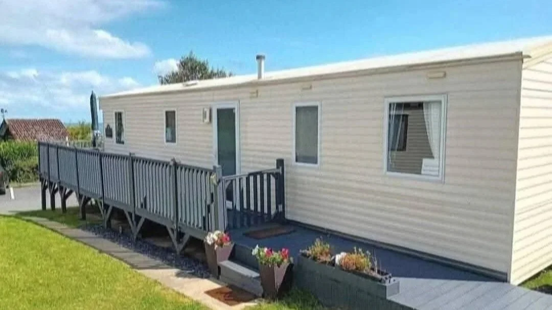 Exterior view of a beige mobile home with white trim, featuring three windows, a sliding glass door, a small wooden deck with a ramp, and flower pots nearby, set in a grassy yard under a partly cloudy sky.