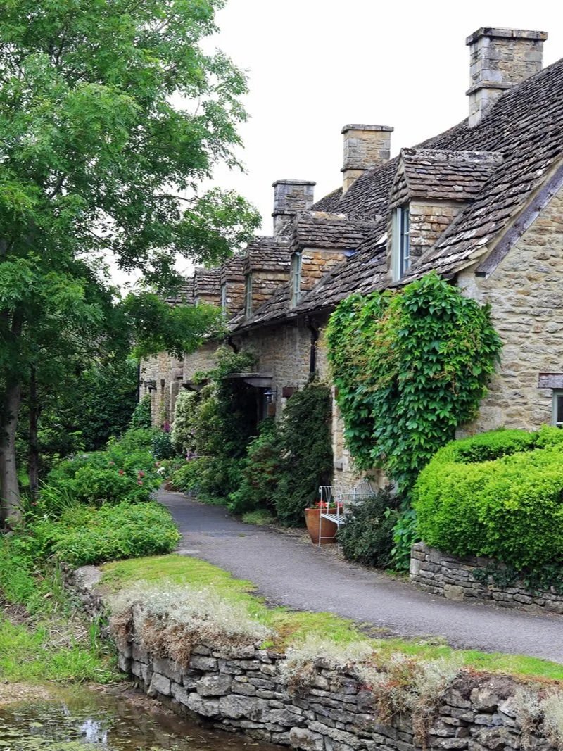 A row of stone cottages with sloped roofs and multiple chimneys, surrounded by lush green bushes and trees along a winding pathway.