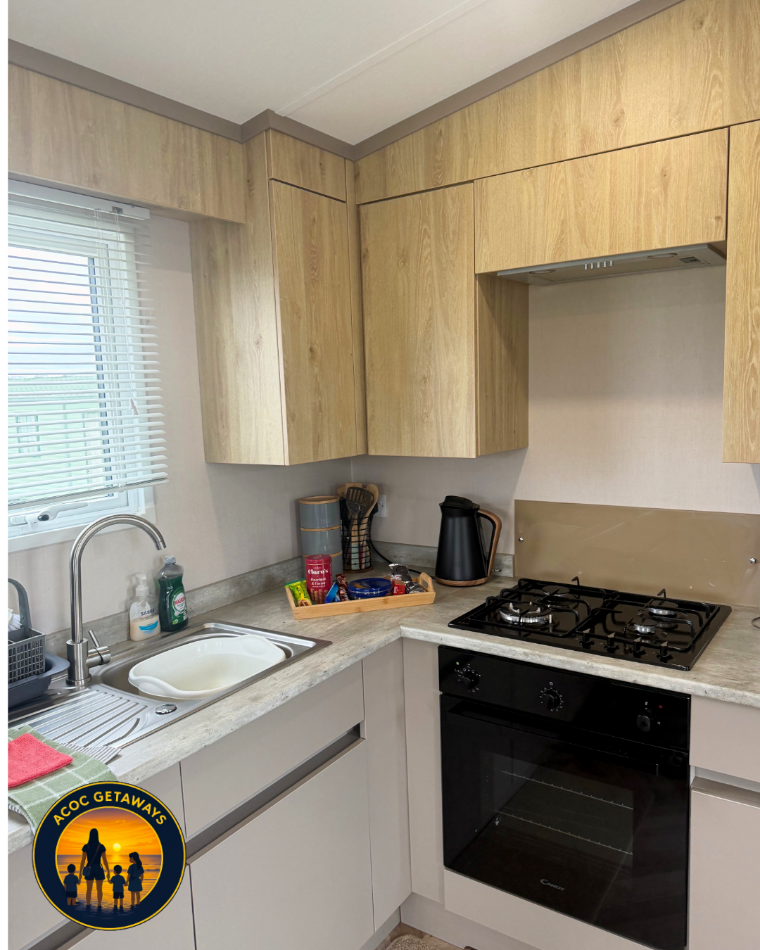 Kitchen with light wood upper cabinets, a window with blinds, a stainless steel sink with green dish soap, a black dish drying rack, and a black stove with oven. Countertop has a wooden tray with snacks, a black electric kettle, and a container with 