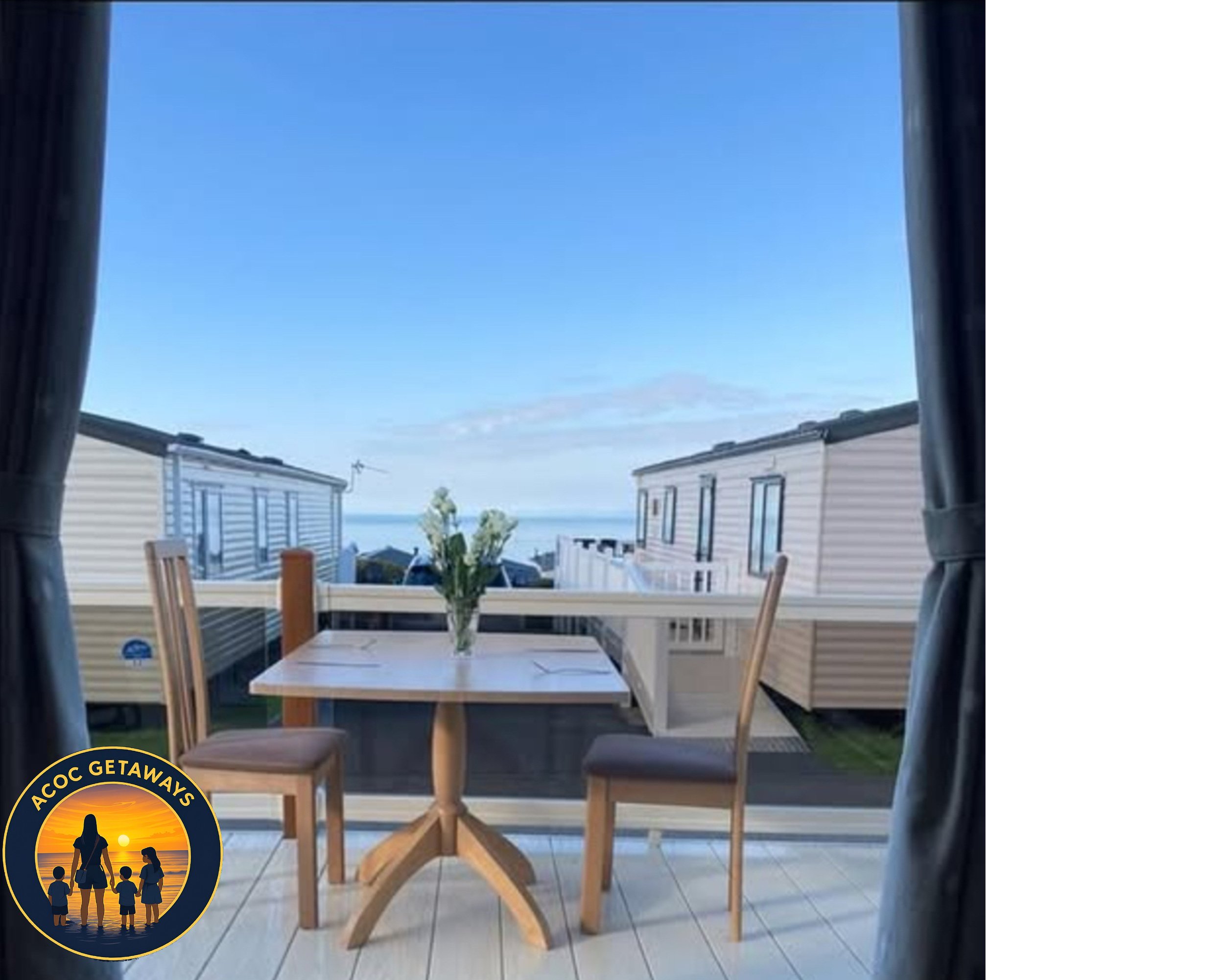 View from a balcony overlooking mobile homes with a wooden table, two chairs, and a vase with flowers, under a clear blue sky.