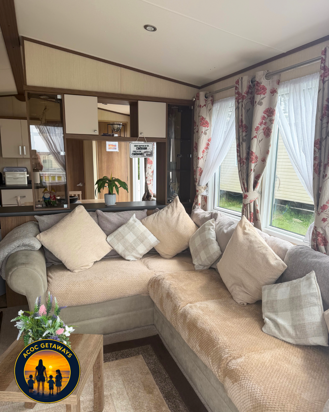 Living room with a beige sectional sofa, filled with beige and checked throw pillows, near windows with floral curtains. A wooden coffee table with a flower arrangement is in front of the sofa. Open kitchen area with a counter visible in the backgrou
