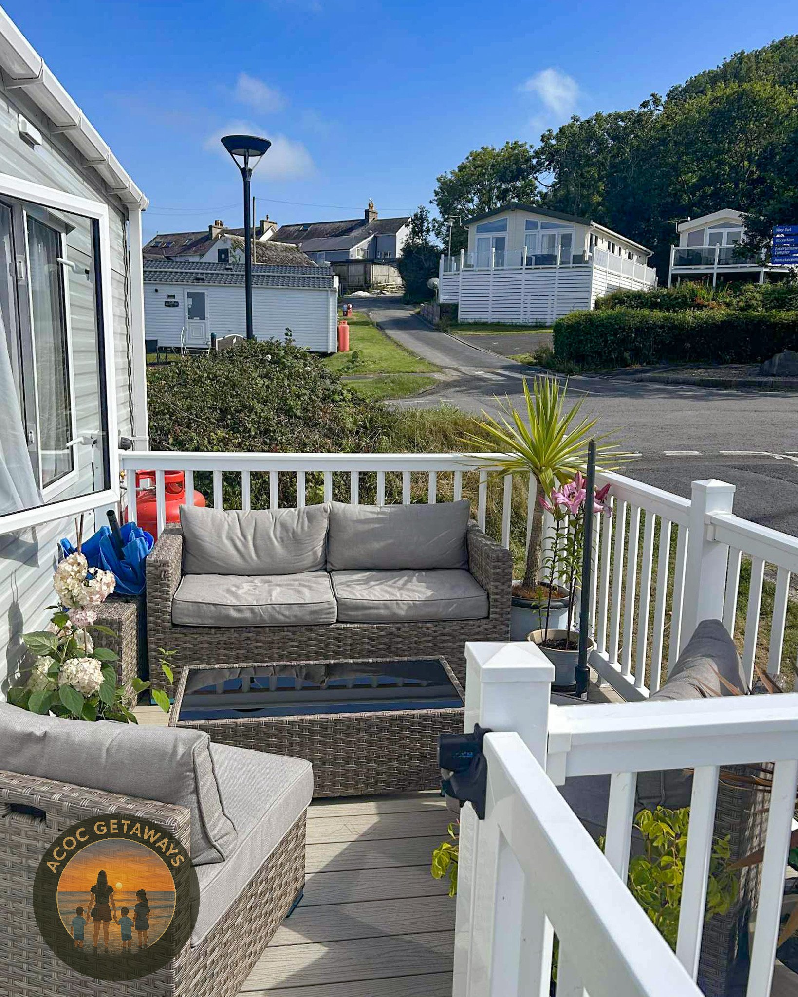 View of a cozy outdoor patio with wicker furniture, potted plants, and a white railing, overlooking a neighborhood with houses and a hill on a sunny day.