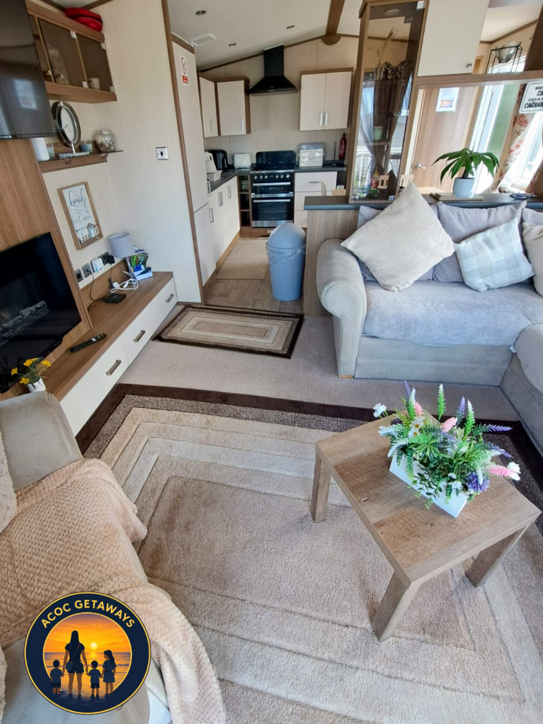 Cozy living room with a beige sofa, a wooden coffee table with a flower arrangement, and a carpeted floor. The open kitchen area with white cabinets, a black stove, and small appliances can be seen in the background. A television with electronics and