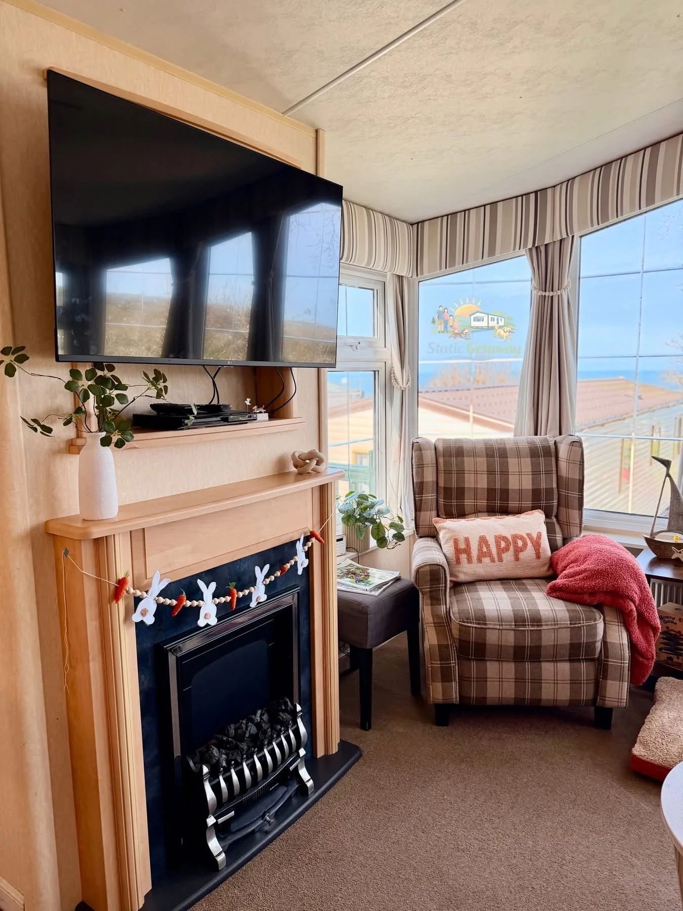 A cozy living room corner with a fireplace decorated with a felt banner, a plaid armchair with a 'HAPPY' pillow and pink blanket, a small side table, and a large window with view of the sky and rooftops, bright daylight.