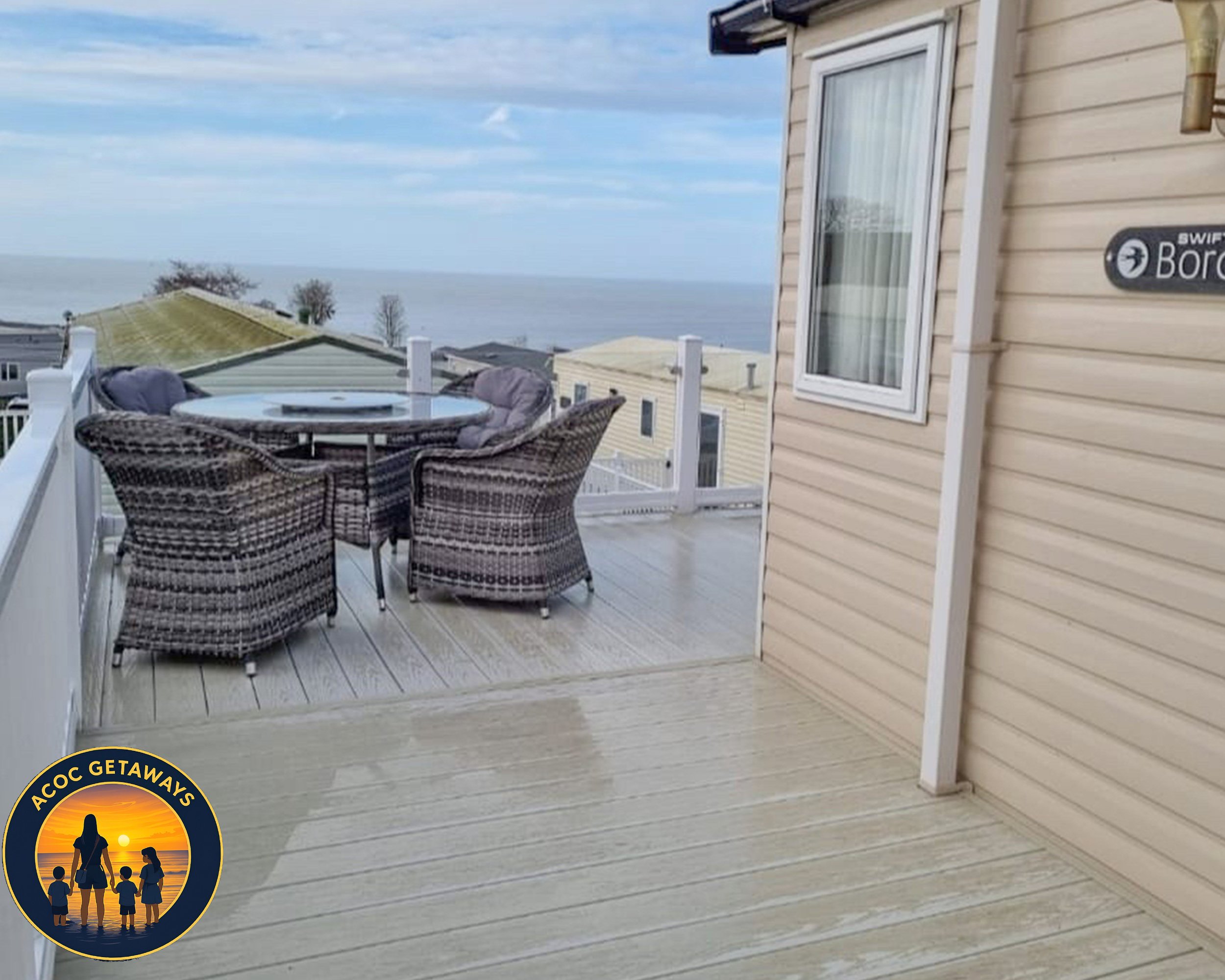 A wooden deck with a round table and four wicker chairs, overlooking a view of neighboring houses, the ocean, and the sky.