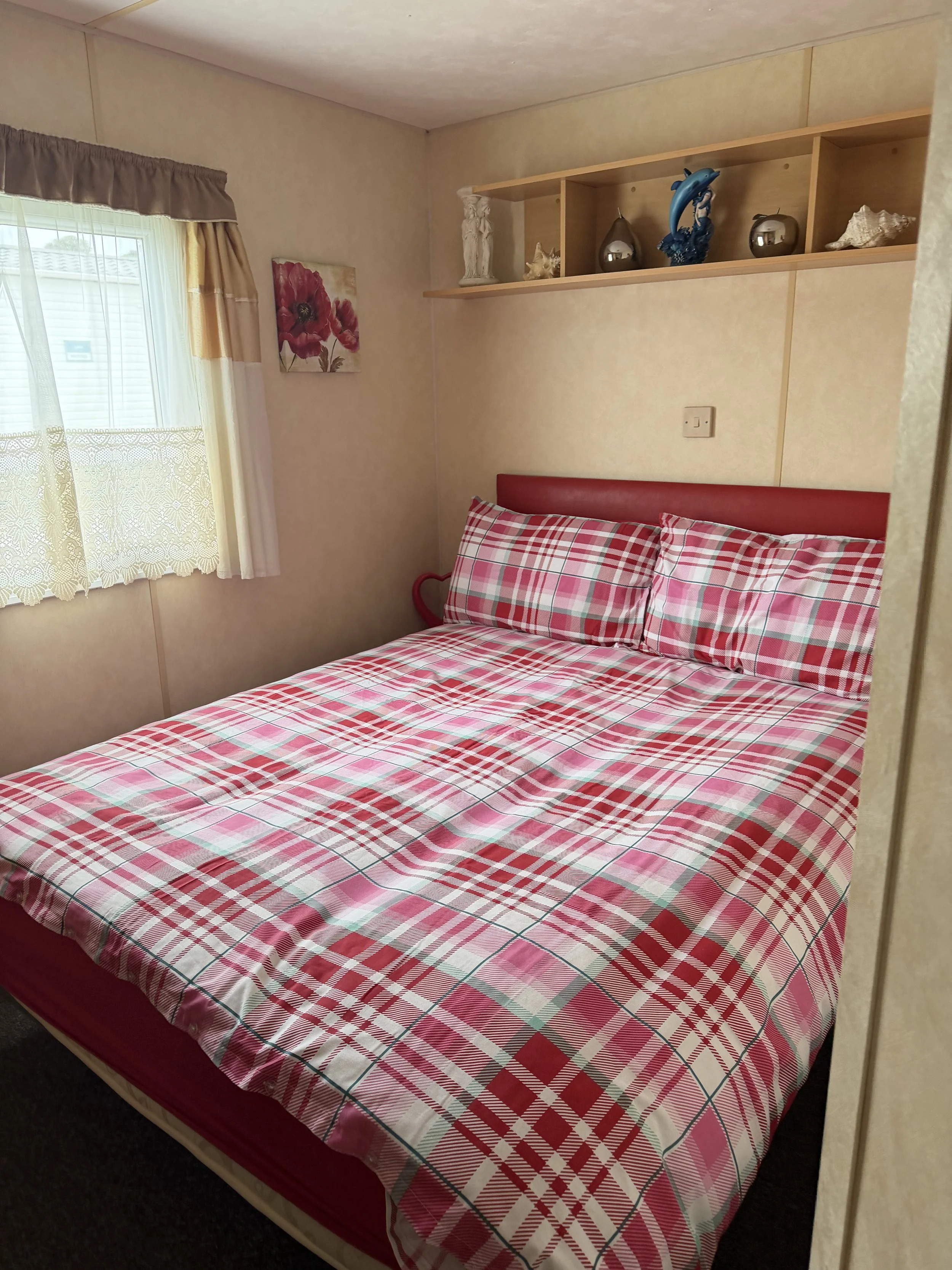 A bedroom with a bed covered in red, white, and green plaid bedding, a window with lace curtains and beige valance, a wall-mounted shelf with decorative items, and a framed floral painting on the wall.