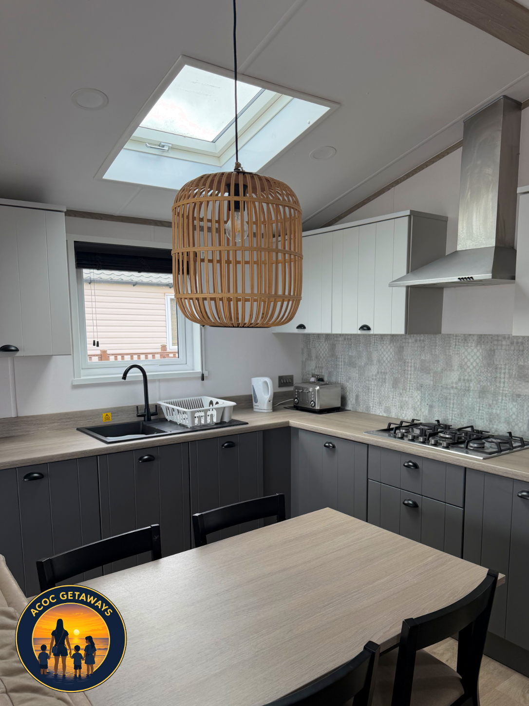 Modern kitchen with white upper cabinets and dark lower cabinets, a skylight, a hanging wooden basket-style light fixture, and a window with black blinds showing an outside house wall. There is a dining table with black chairs, a white dish rack, ket
