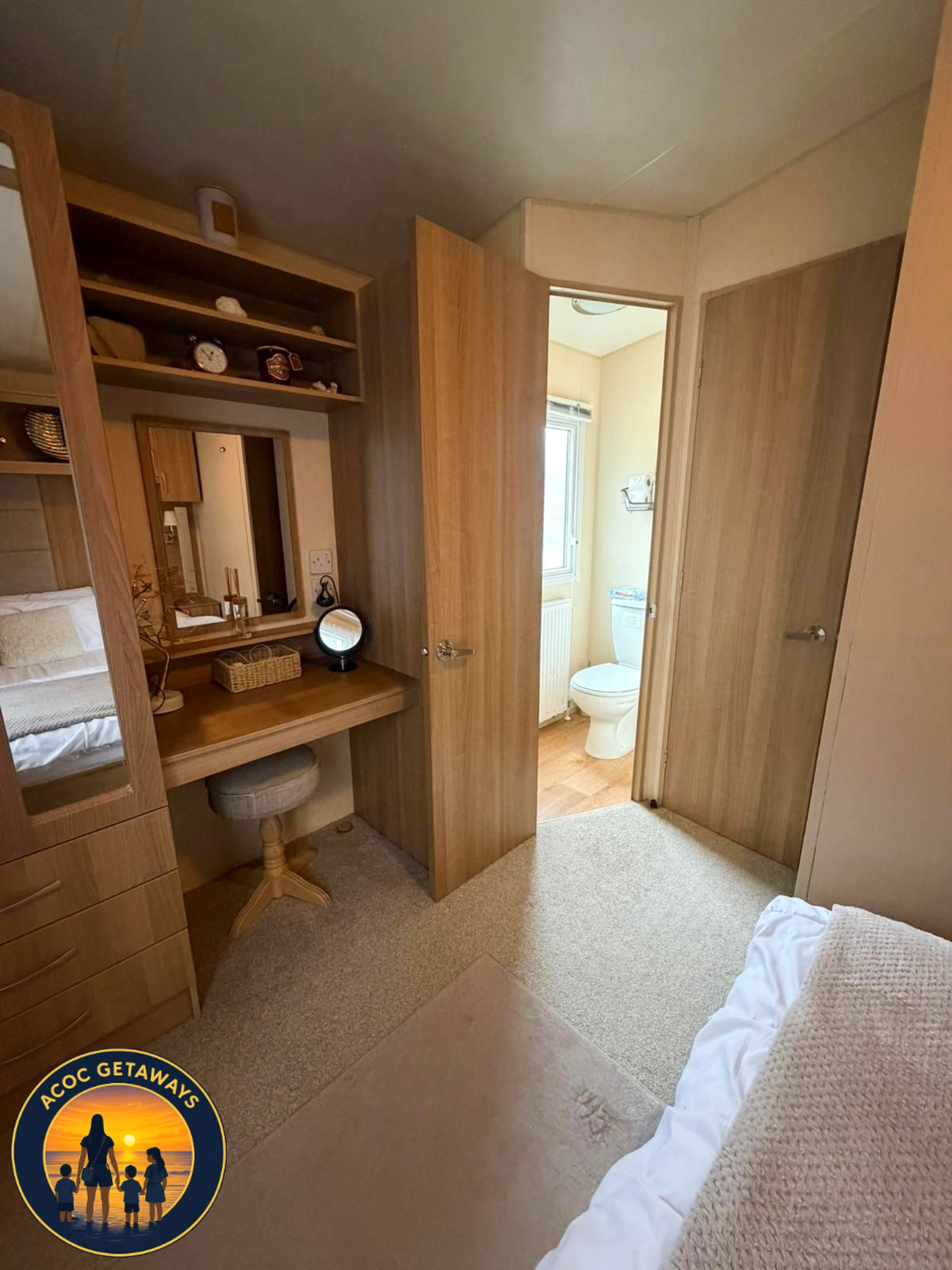 View of a cozy bedroom with a wooden vanity, mirror, and a stool. An open door reveals a small bathroom with a toilet and window.