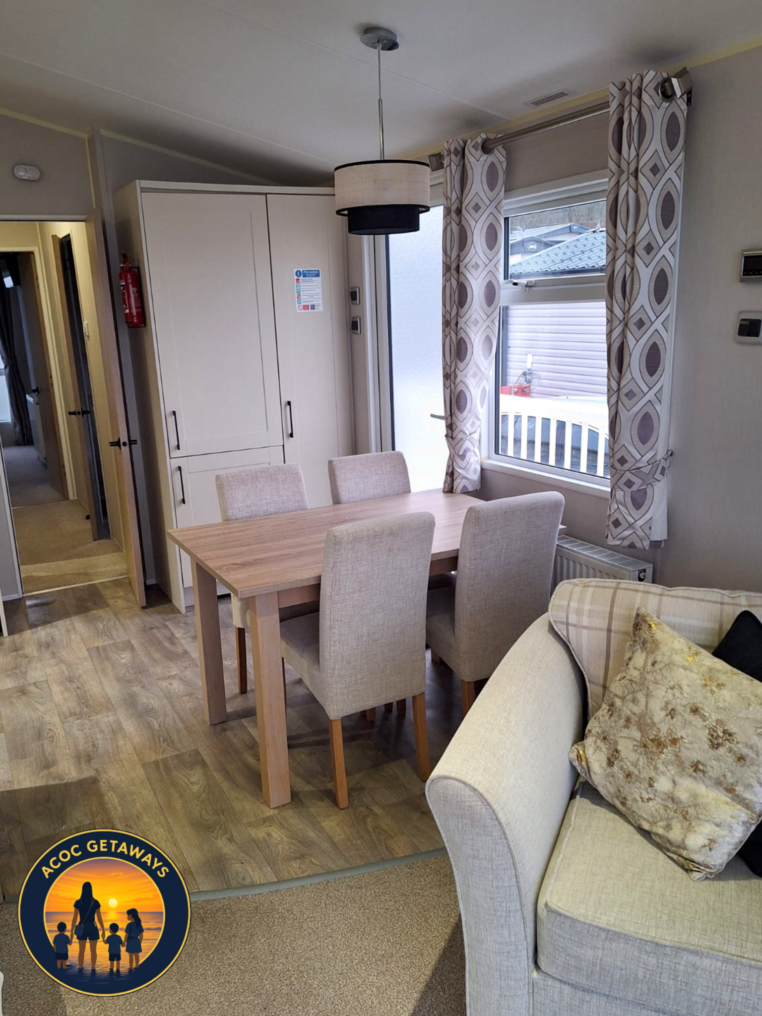 Interior of a cozy living and dining area with a light-colored sofa, a beige pillow, a wooden dining table, beige upholstered chairs, a white cabinet, a window with geometric patterned curtains, and a ceiling light fixture.