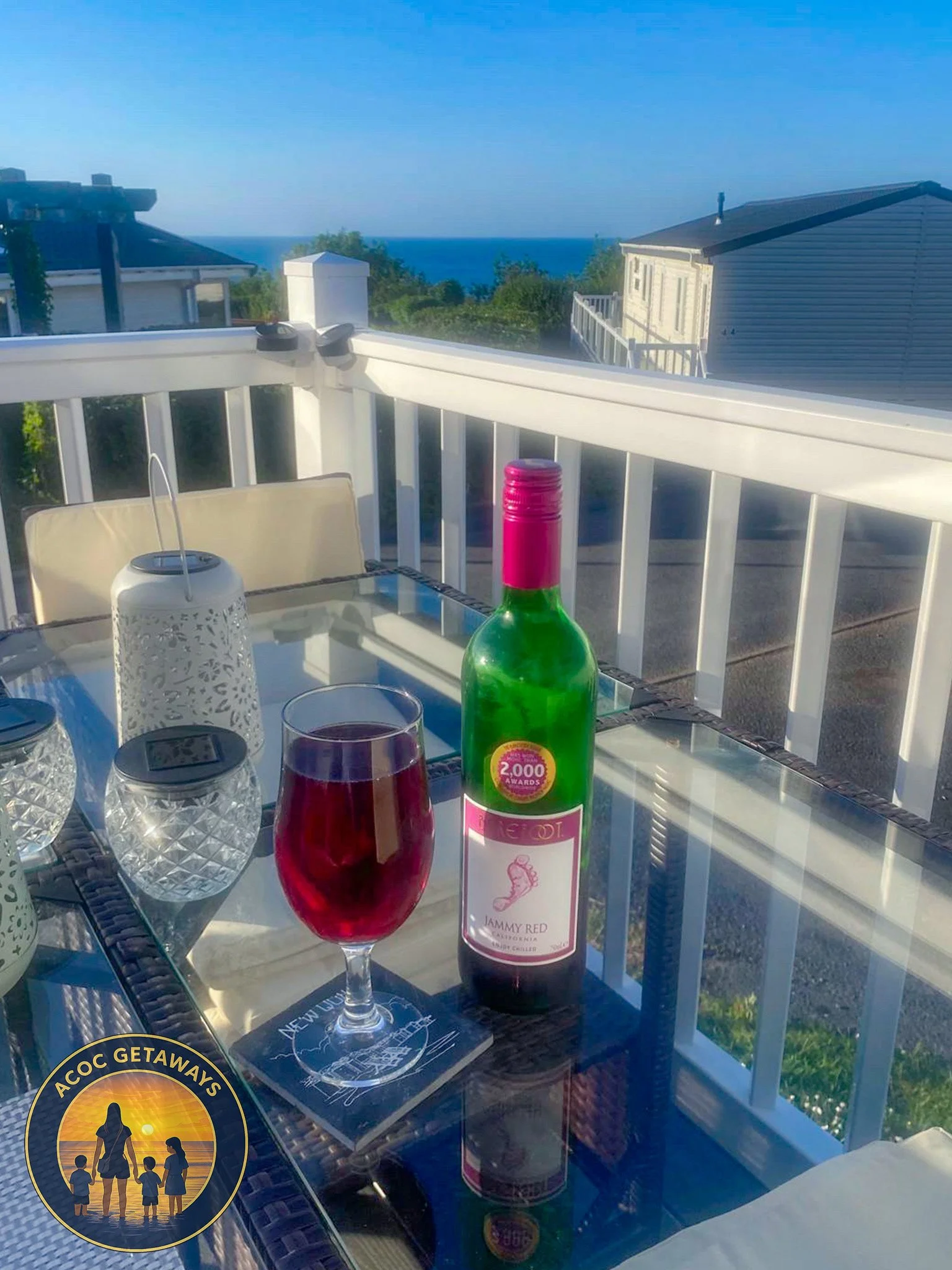 Bottle of Merlot wine, glass of red wine, two lidded glasses, and a decorative lantern on a glass outdoor table, with a ocean view background on a sunny day.