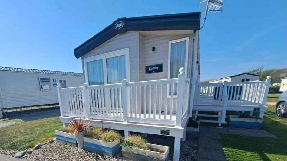 White tiny house with deck and railing, located in a trailer park, under a clear blue sky.