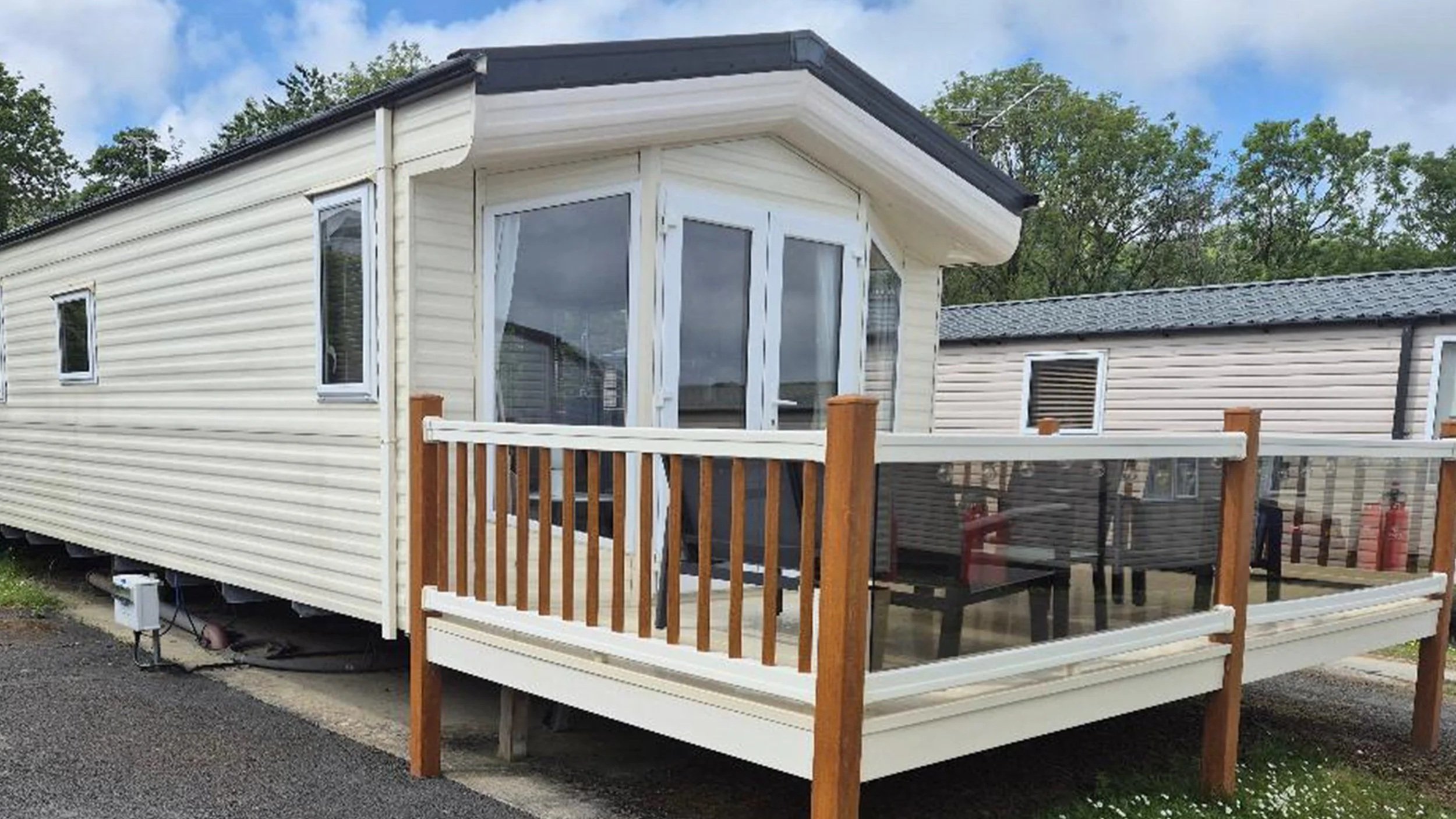 A white mobile home with a small outdoor deck featuring wooden posts, a railing with glass panels, and a sliding door. The mobile home has three small windows and is situated on a gravel lot with trees in the background.