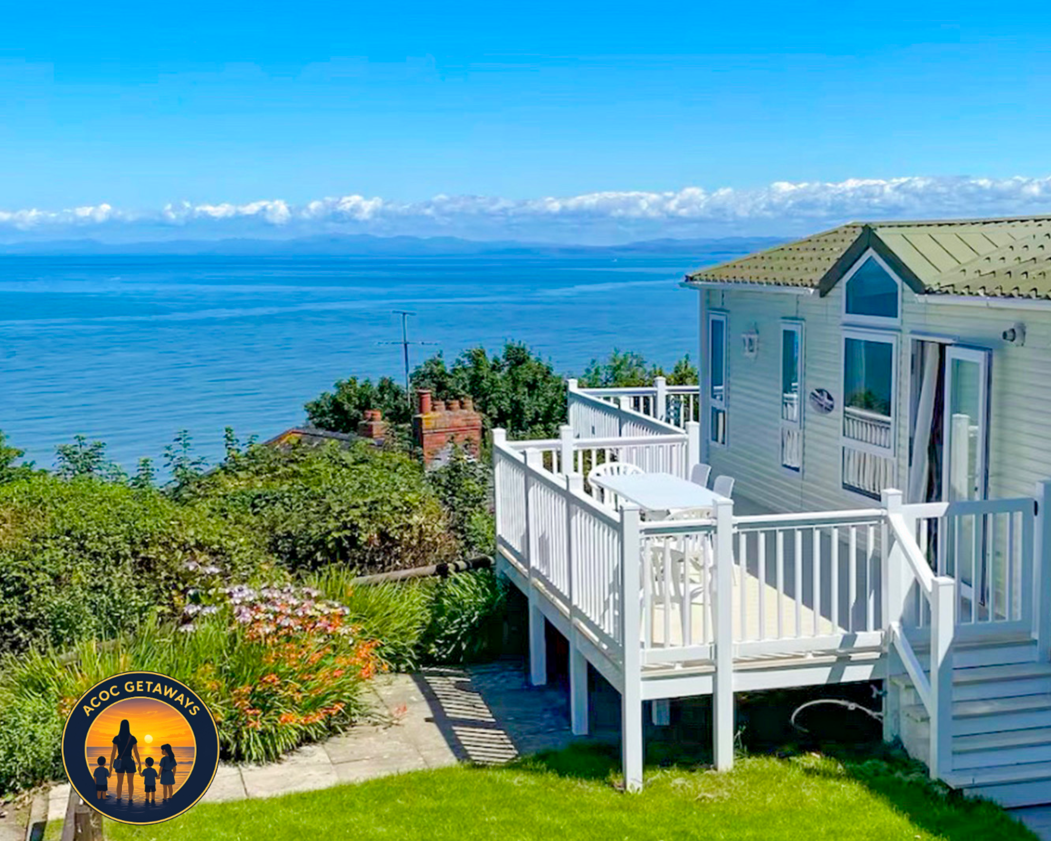 A coastal house with a green roof and white balcony, overlooking a calm blue sea under a partly cloudy sky, with lush garden plants in the foreground.