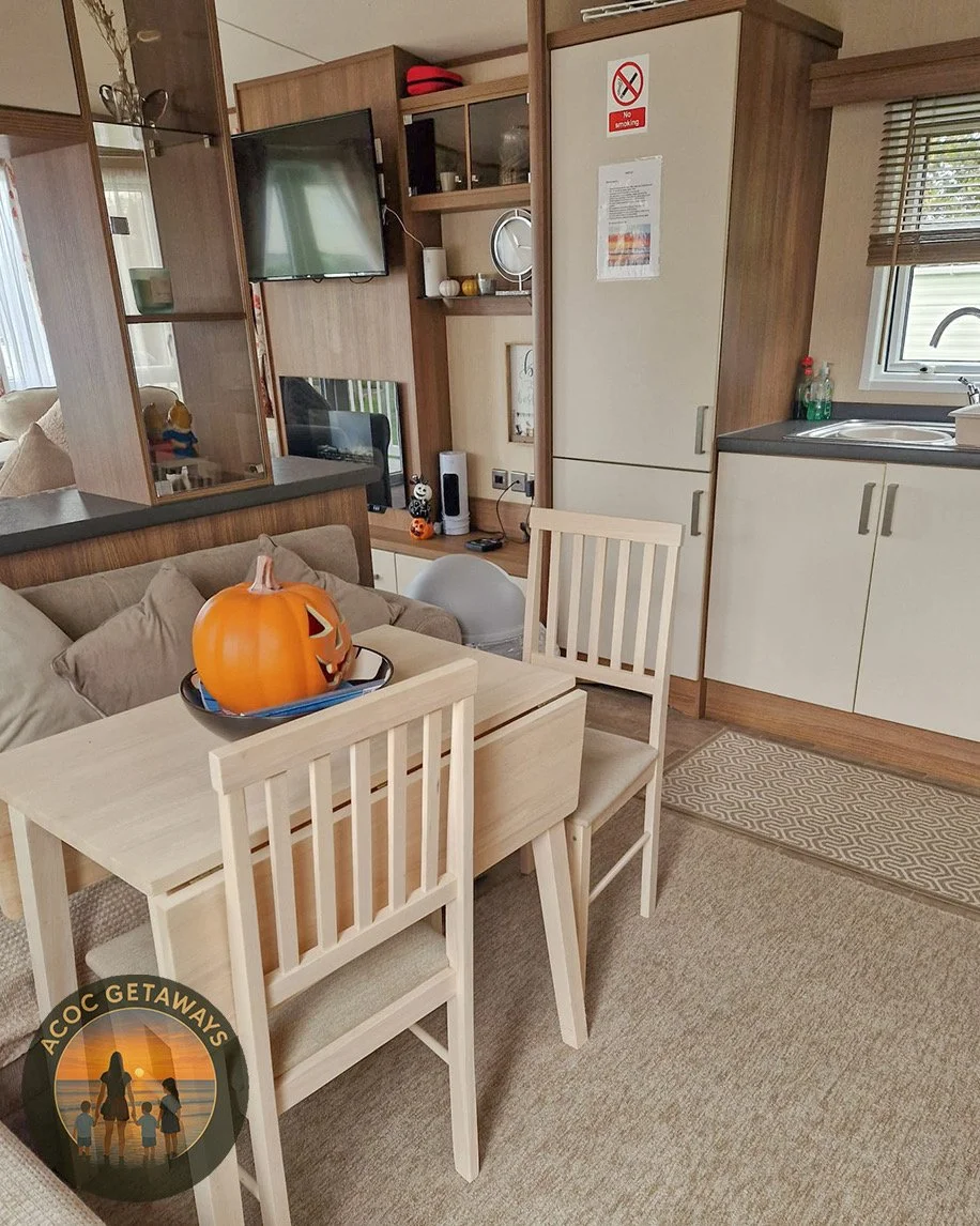 Interior of a cozy living area with a dining table decorated for Halloween, featuring a pumpkin-shaped container with a carved face. The space includes a kitchen with a sink, window, and cabinets, along with a small entertainment area with a TV and decorative items.