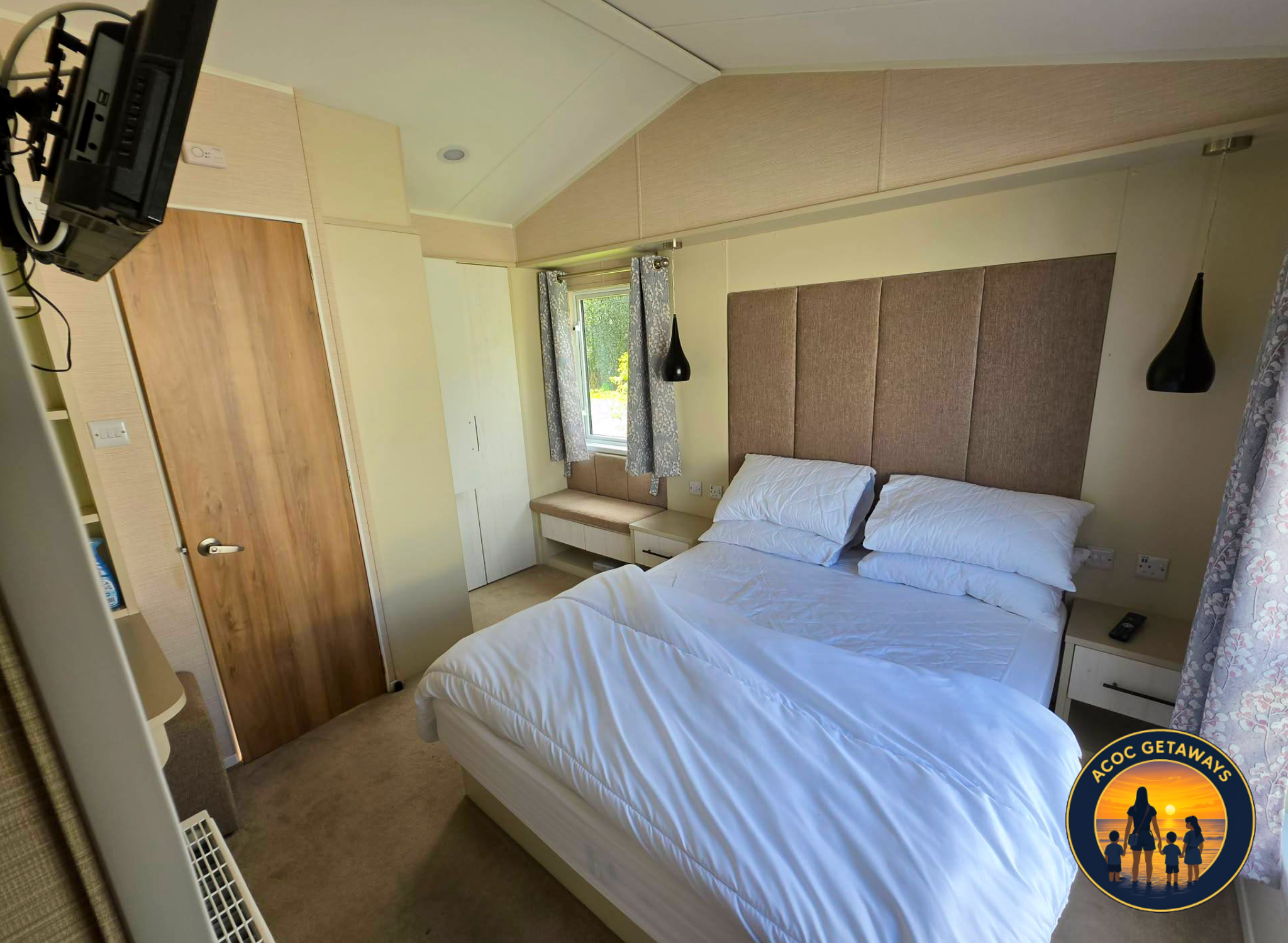 A bedroom with two single beds, beige carpet, one nightstand with a remote control, a window with curtains, hanging black pendant lights, and a wooden chest of drawers with an electronic device on top.