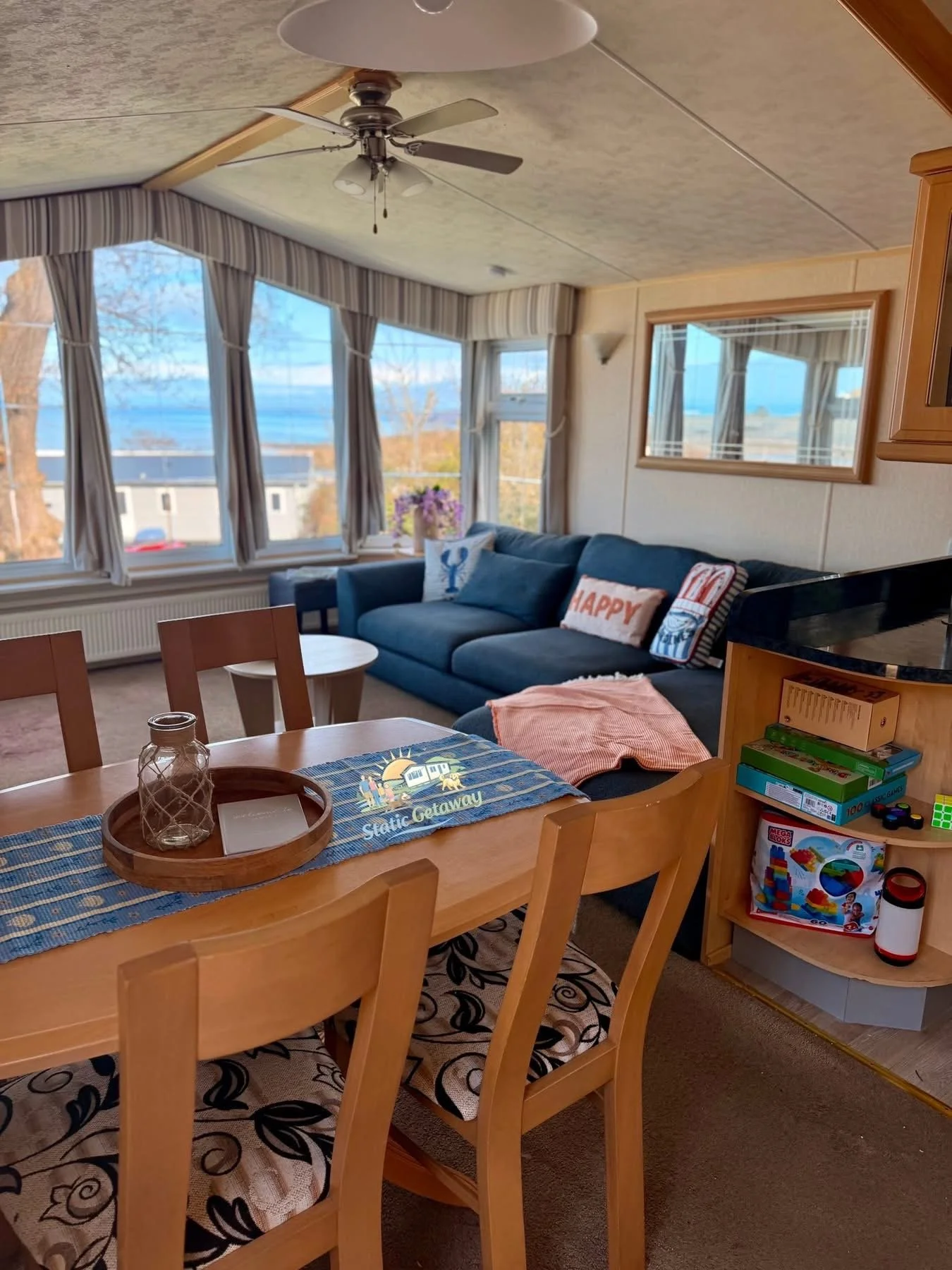Living room with large windows showing a view of the ocean, with a blue couch, cushions, a glass vase on a table, and a ceiling fan.