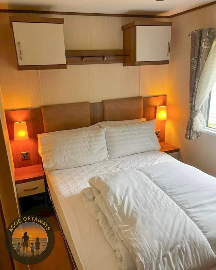 A cozy bedroom with a bed, two white pillows, and a white comforter. There are wall-mounted night lamps on each side of the bed. A window with curtains is on the right, allowing natural light into the room. Above the bed is a wooden storage unit with cabinets and a shelf.