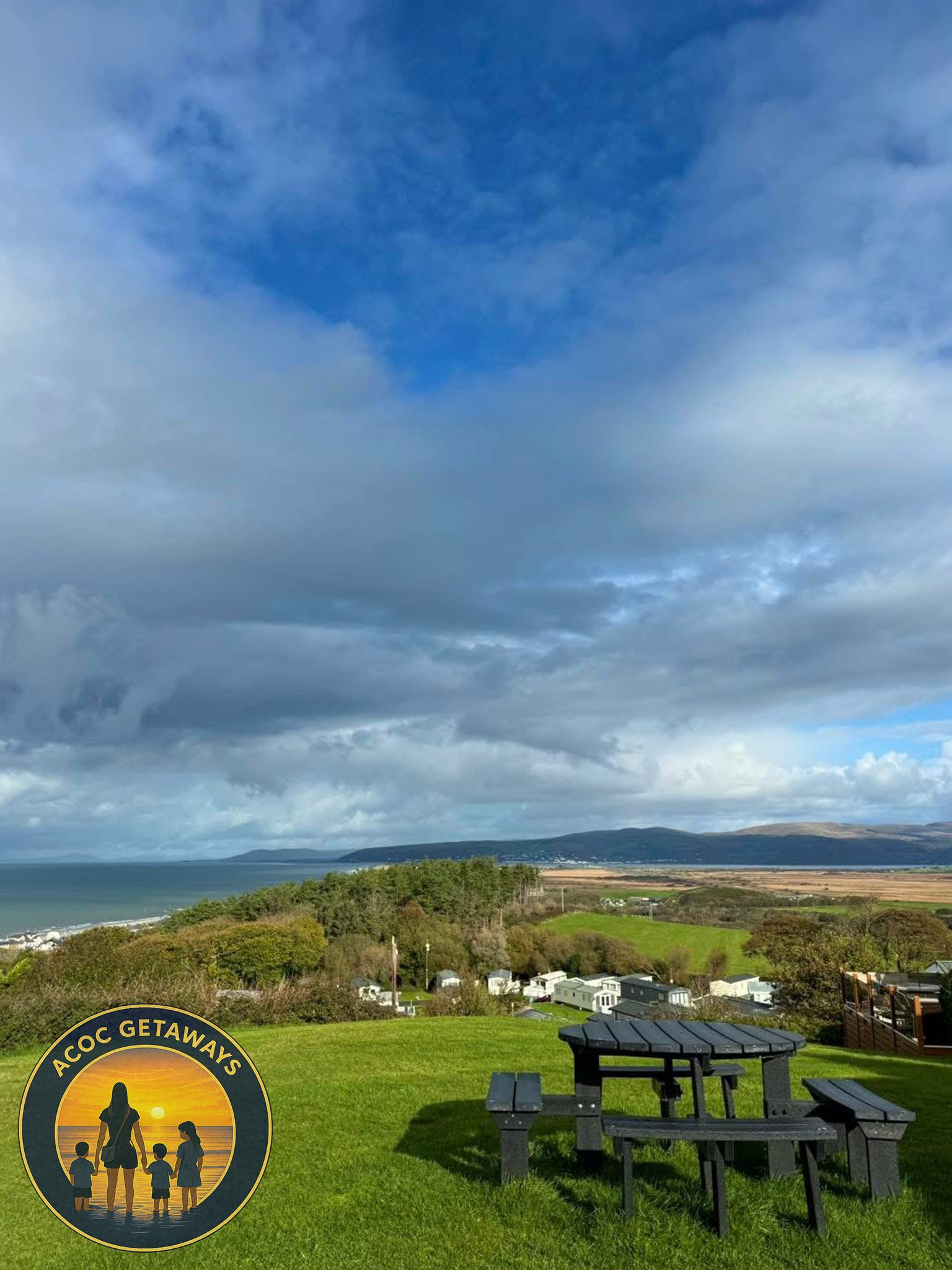 Scenic view of a grassy outdoor area with a black picnic table, overlooking a coastal landscape with hills, a beach, ocean, and partly cloudy sky.
