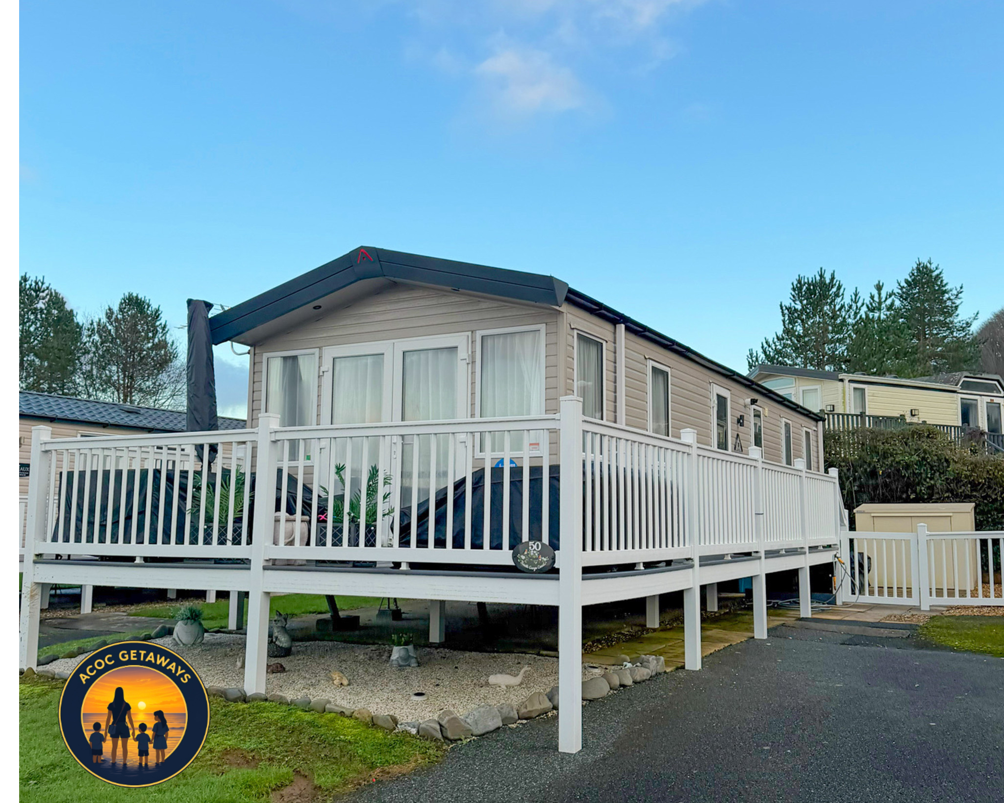 A two-story house with beige siding, multiple large windows with white curtains, and a black roof under a clear blue sky with some clouds. The house has a white deck with a railing and a small garden area underneath with rocks and grass. There are ta