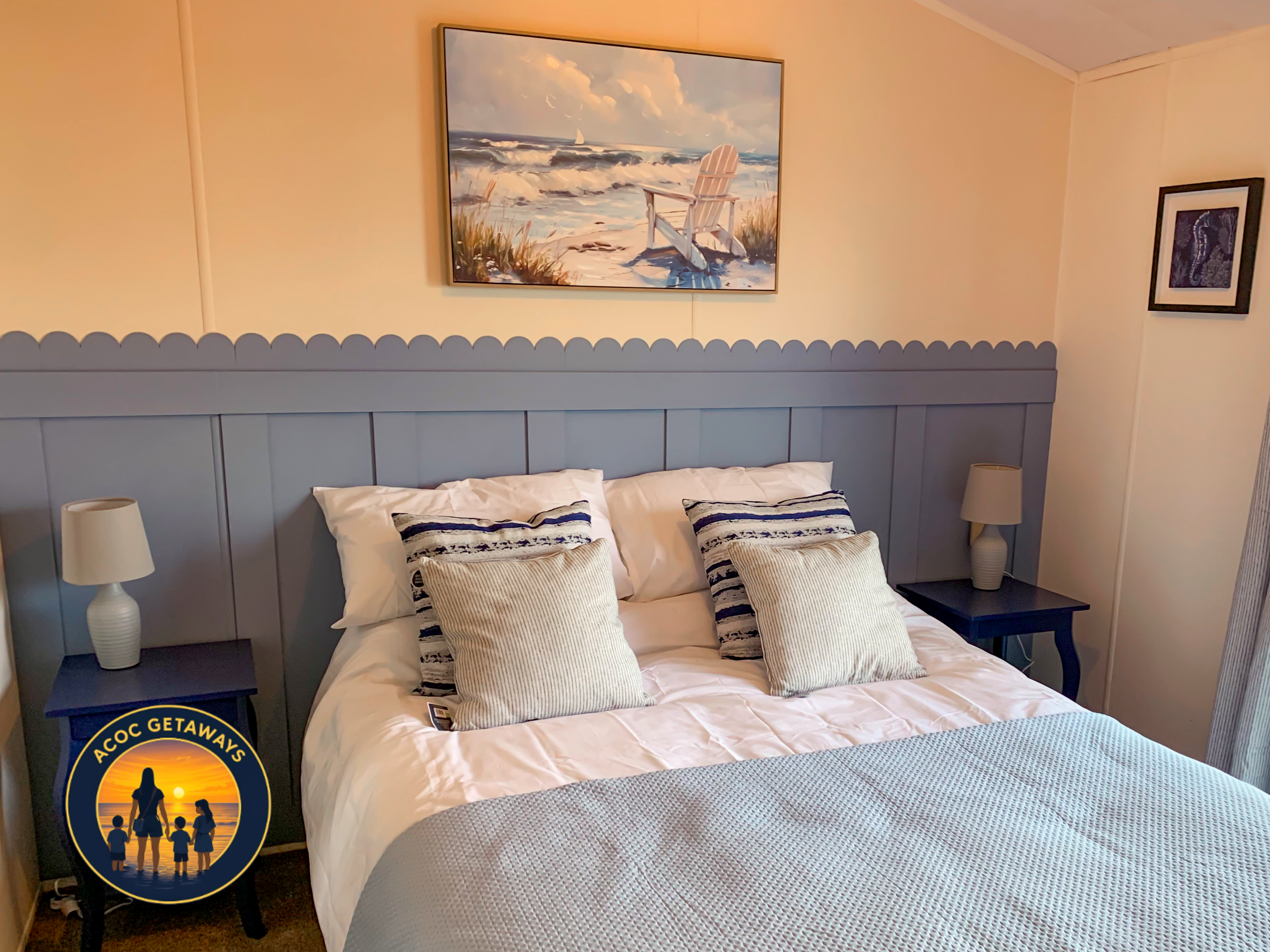A cozy bedroom with a bed covered in white and blue bedding, two pillows with striped covers, and two beige pillows. There are two matching nightstands with lamps on each side of the bed, a wall art of a beach scene with a chair and ocean view above 