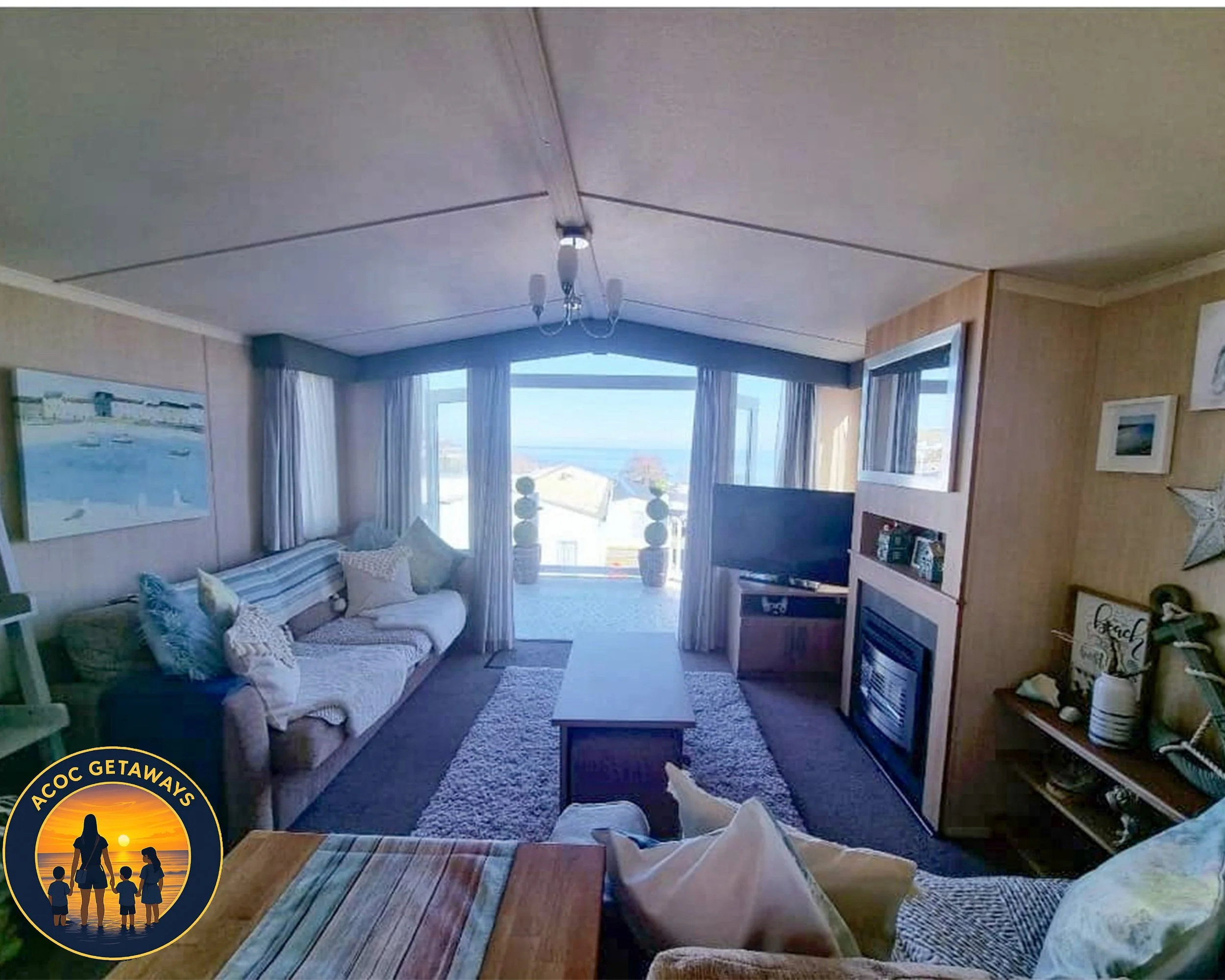 Living room with large windows showing ocean view, beige couch with pillows, TV, fireplace, and dining table.