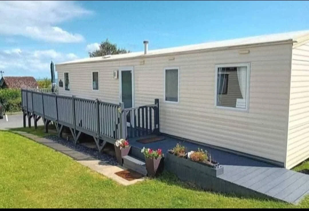 A beige mobile home with a wooden deck and ramp, surrounded by a green lawn and a few potted plants, under a cloudy blue sky.